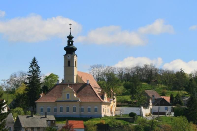 Church with tower in a rural setting, surrounded by trees and houses.