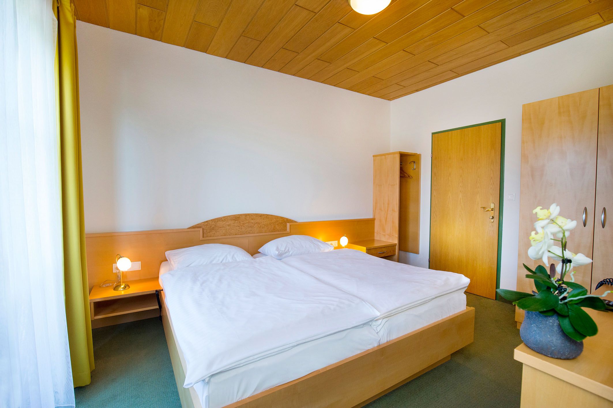 Ein einfach eingerichtetes Hotelzimmer mit Doppelbett, Holzdecke, Schrank und Nachttischlampen.