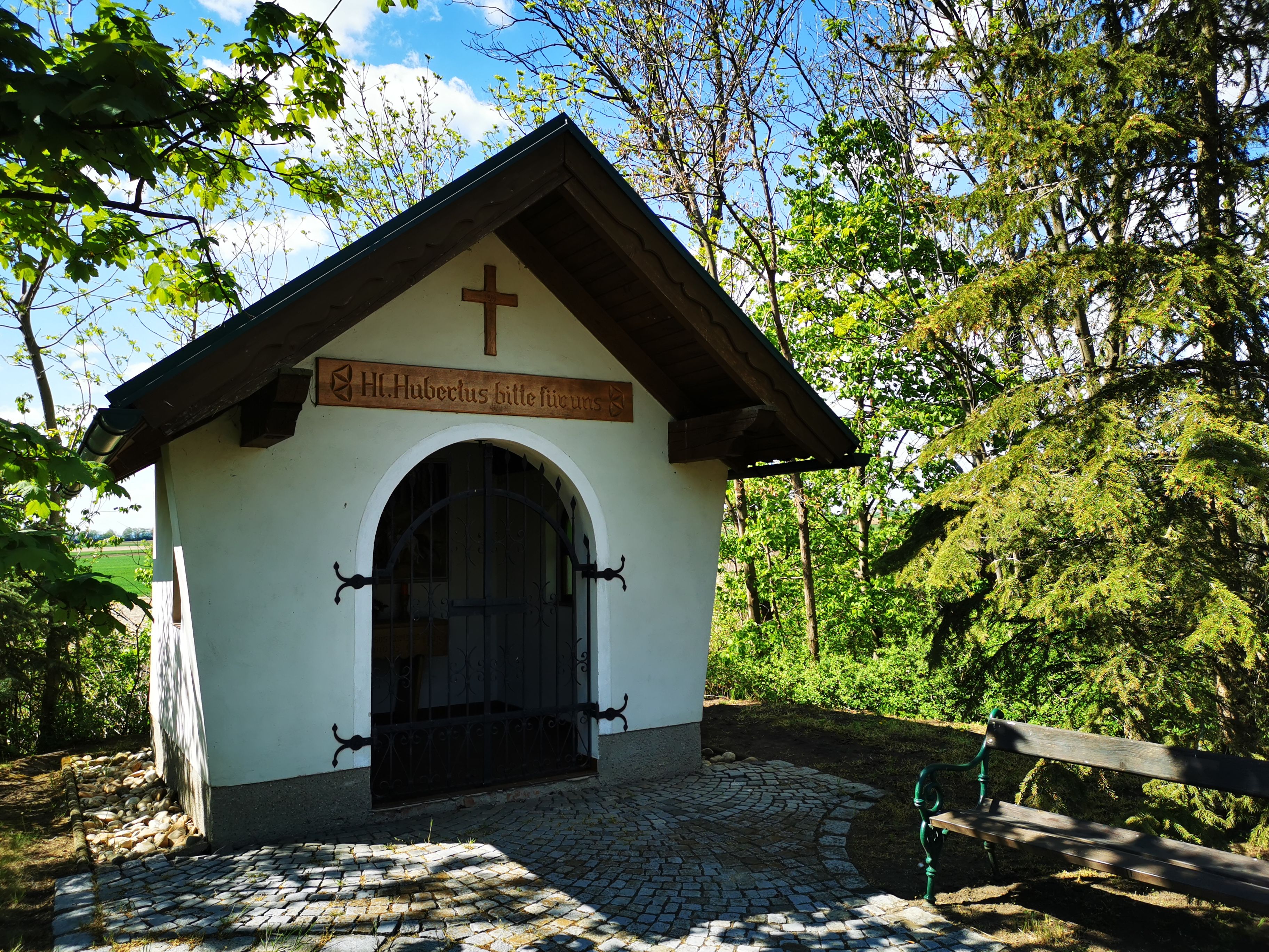 Kleine Kapelle im Grünen mit Holzkreuz und Bank daneben.