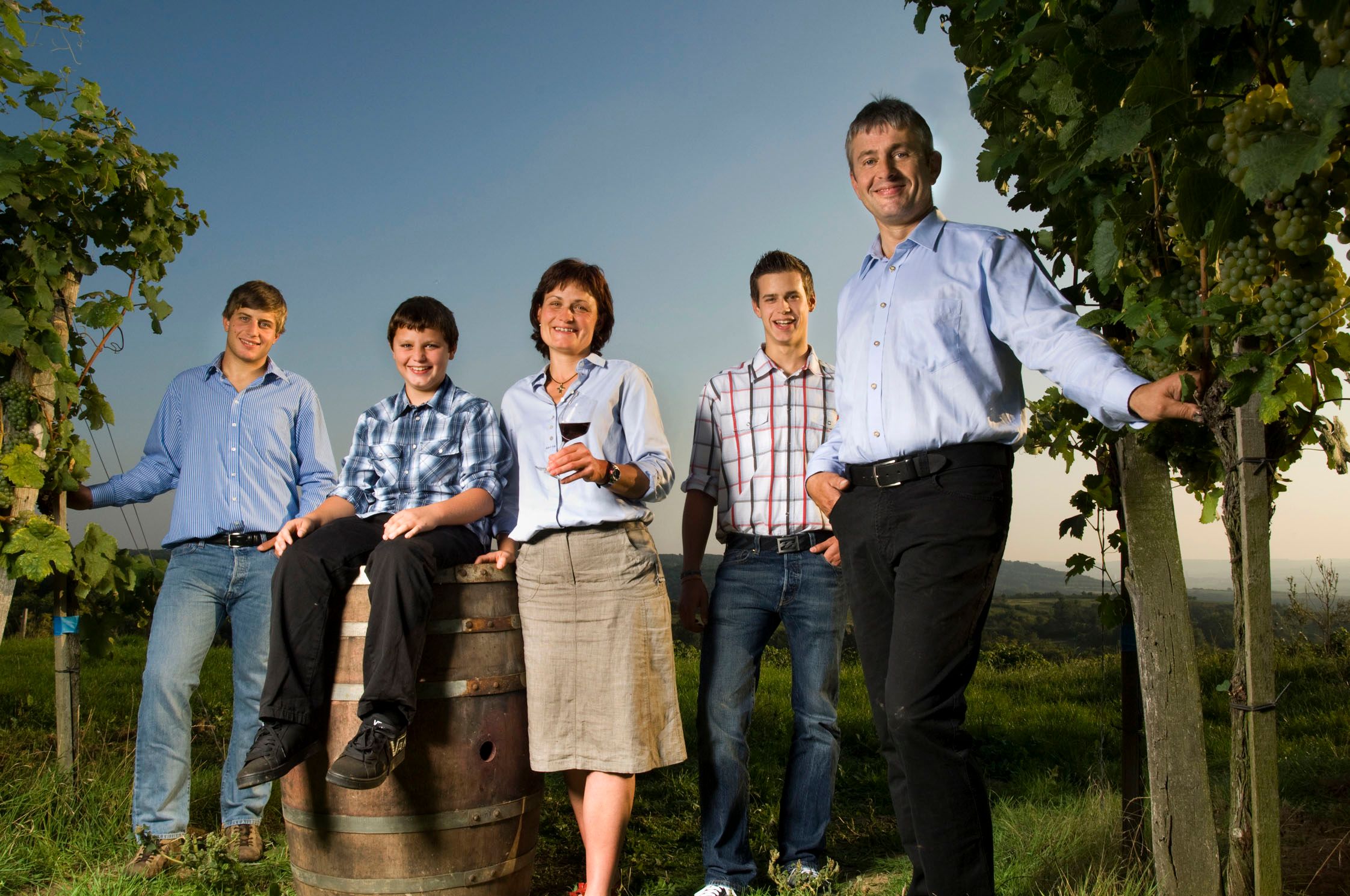A family stands in a vineyard, smiling at the camera. A boy sits on a wine barrel, a woman holds a glass of wine.