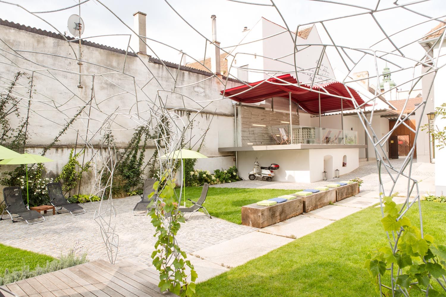 Moderner Innenhof mit Rasen, Liegestühlen, Metallskulpturen und einer Vespa unter einem roten Vordach.