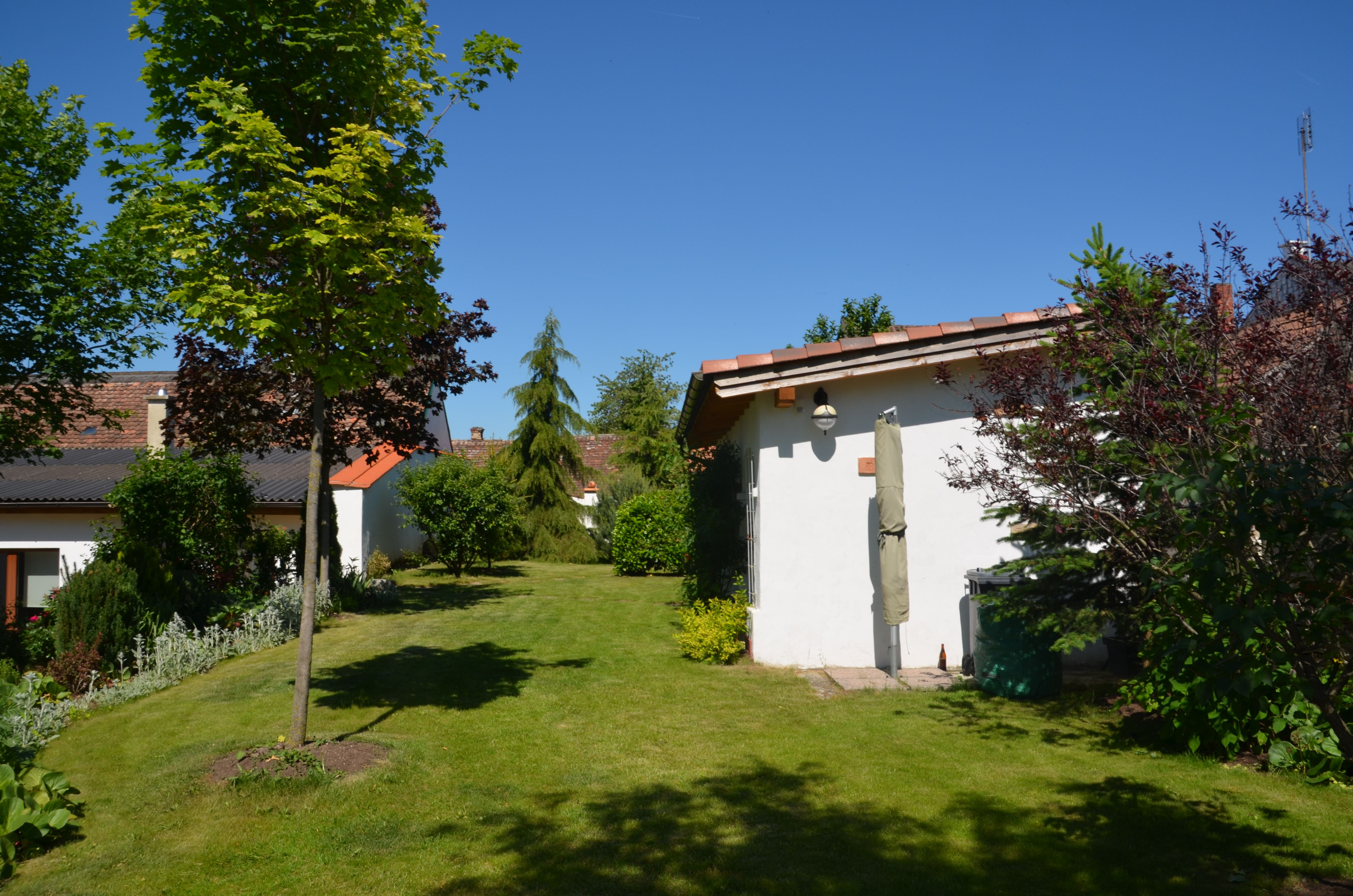 A well-tended garden with lawn, trees and shrubs. Two small buildings with red roofs can be seen in the background.