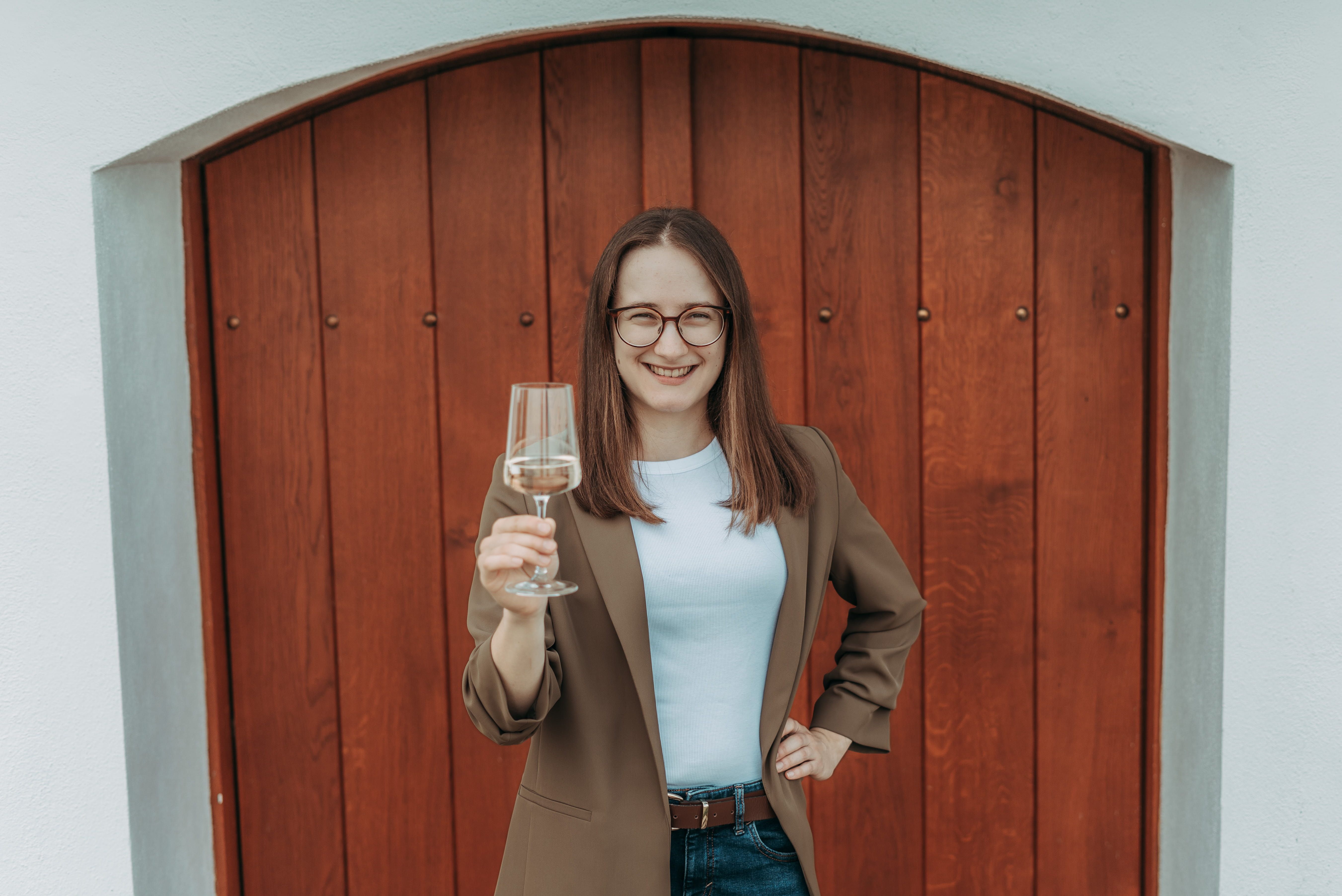 Eine Frau mit Brille hält ein Weinglas und lächelt vor einer Holztür.