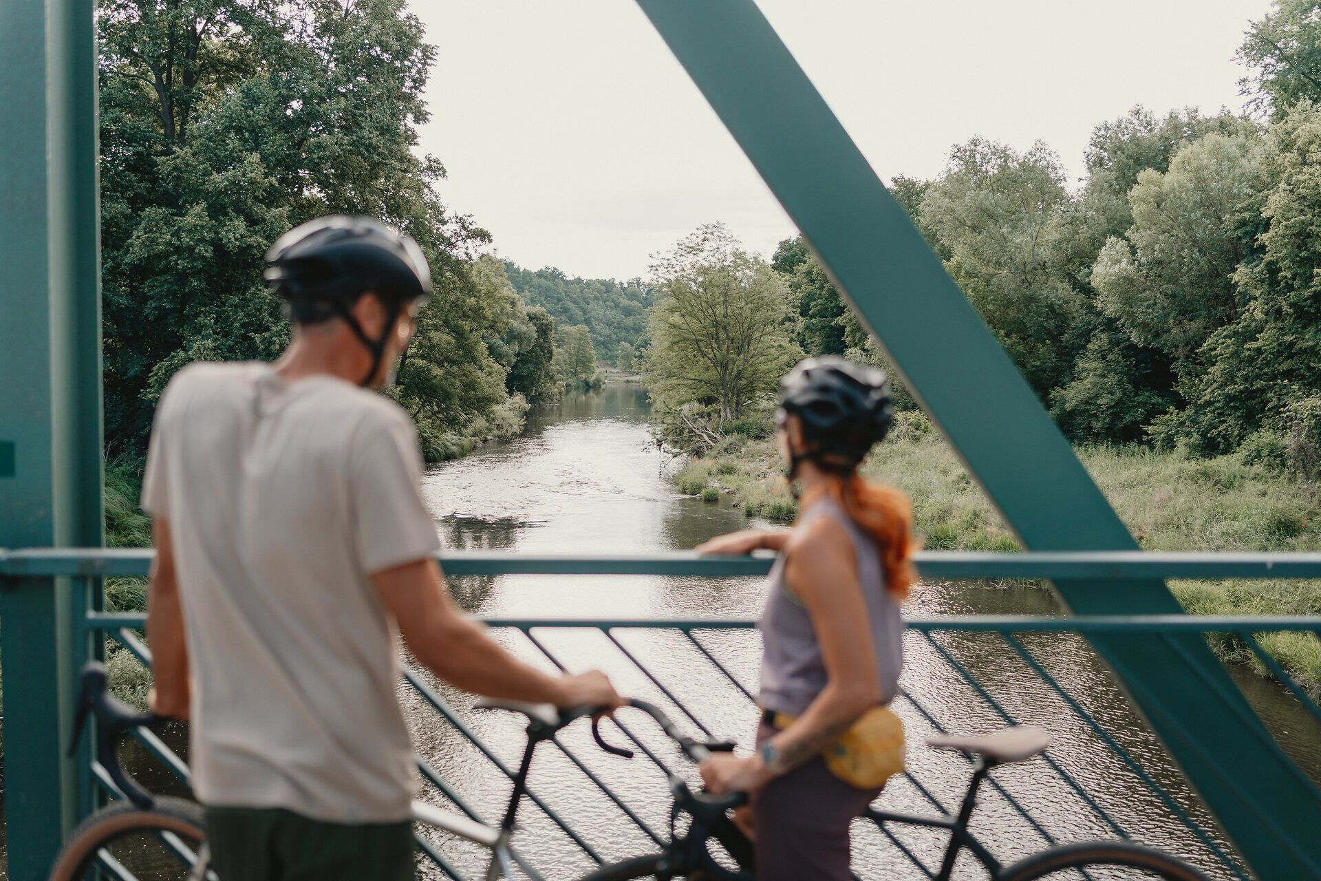 Zwei Radfahrer mit Helmen stehen auf einer Brücke und blicken auf einen Fluss, umgeben von üppiger Vegetation.