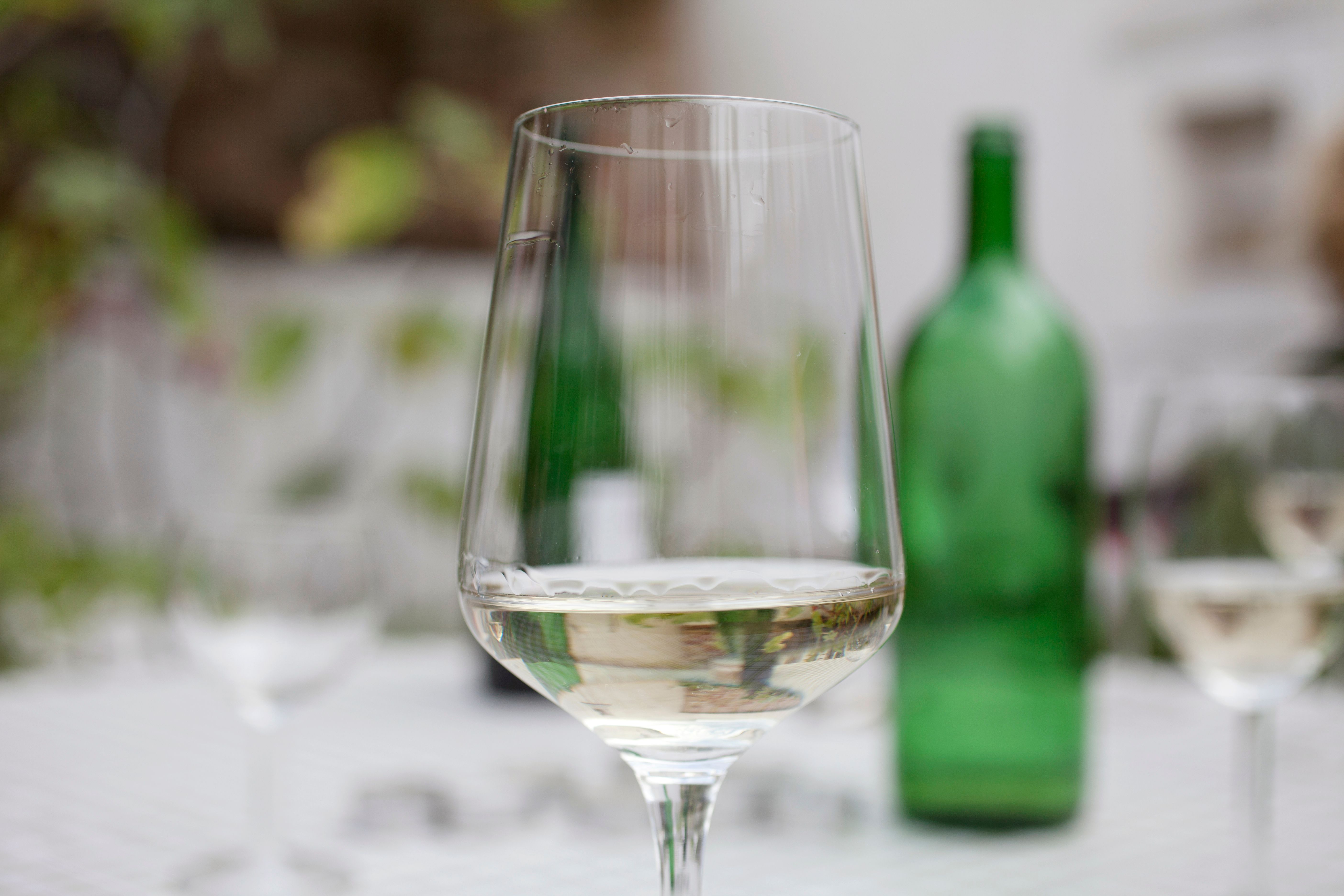 A wine glass with white wine in the foreground, blurred green bottle in the background.