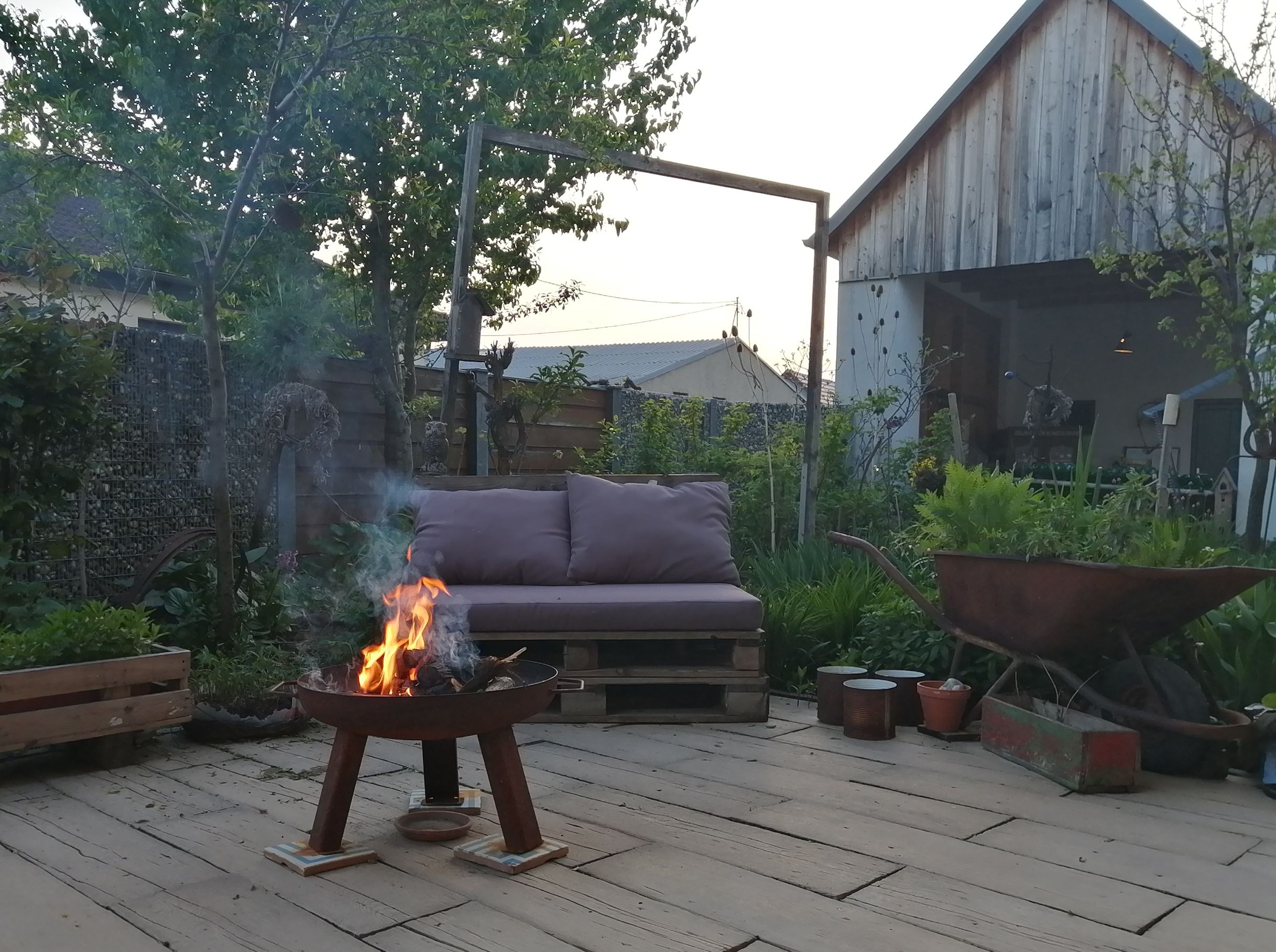 Ein gemütlicher Innenhof mit Feuerstelle, Sofa aus Paletten und einer alten Schubkarre. Im Hintergrund ein Holzschuppen und grüne Pflanzen.