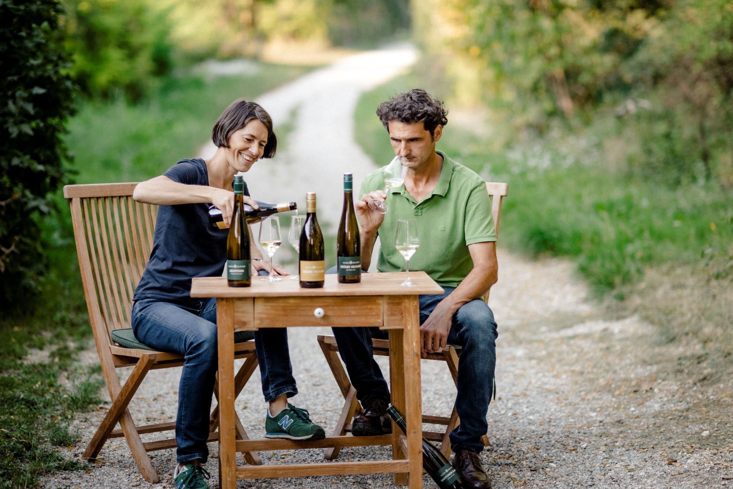 Two people sit at a wooden table outside and taste wine.