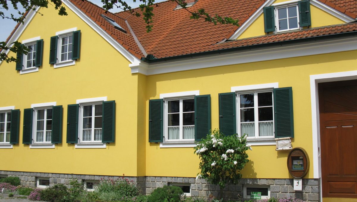Žltý dom so zelenými okenicami a červenou škridlovou strechou.