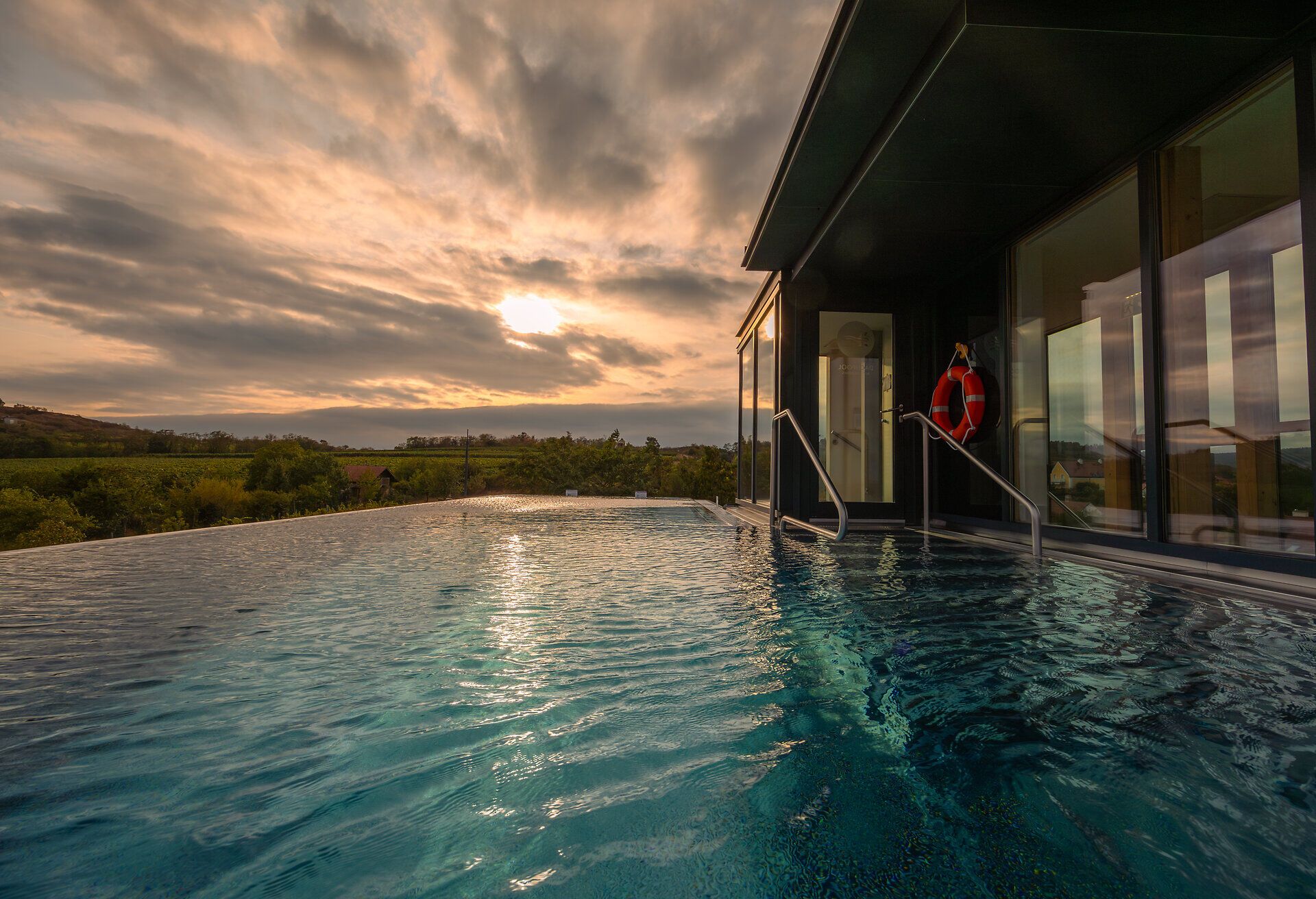 Ein atemberaubender Blick auf den Infinity-Pool, der sanft in die umliegende Landschaft übergeht. Die Wolken am Himmel reflektieren die warmen Farben des Sonnenuntergangs und schaffen eine friedliche Atmosphäre, die zum Entspannen einlädt.