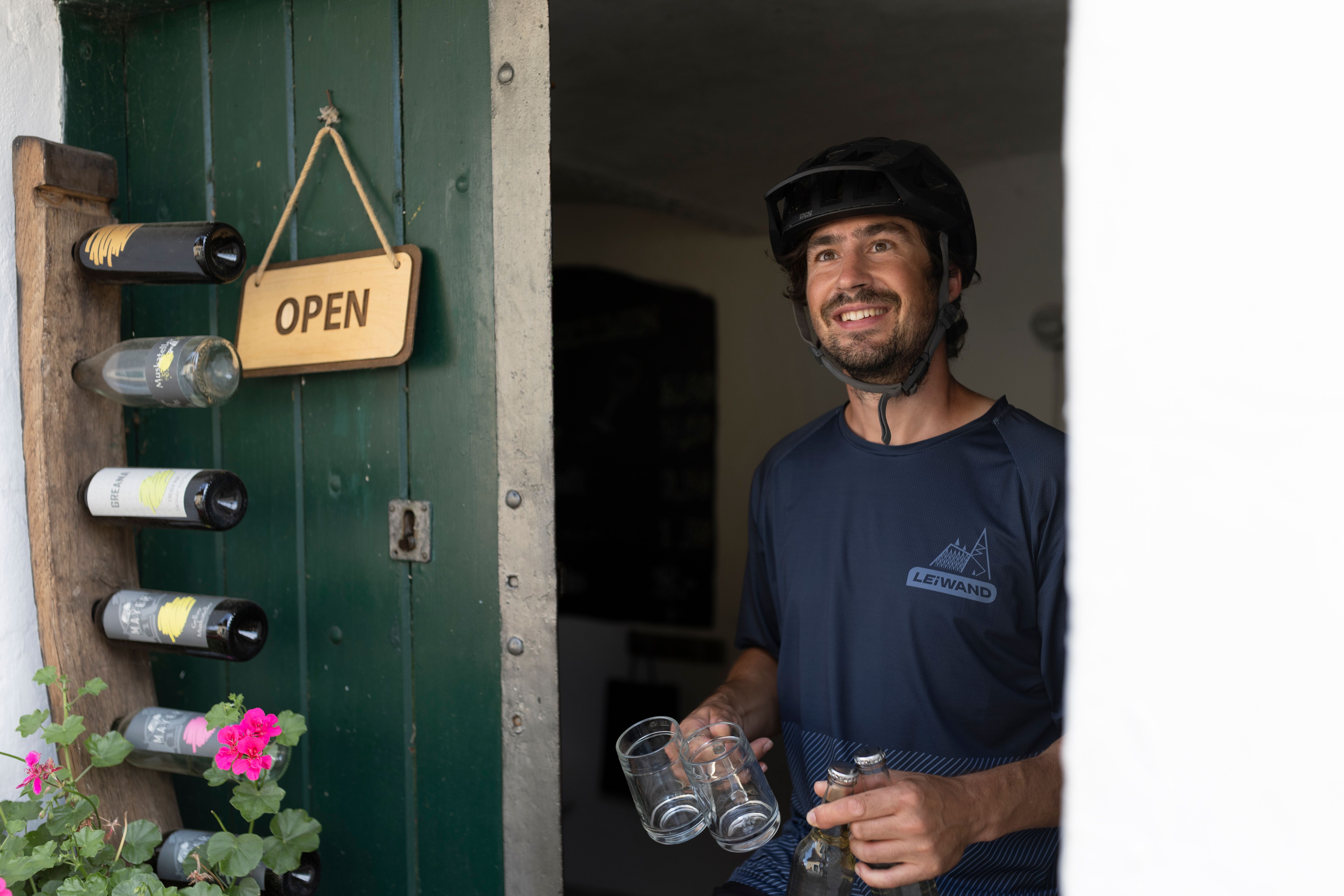A man with a bicycle helmet stands smiling in an open doorway, holding glasses and bottles. An "Open" sign hangs on the door.