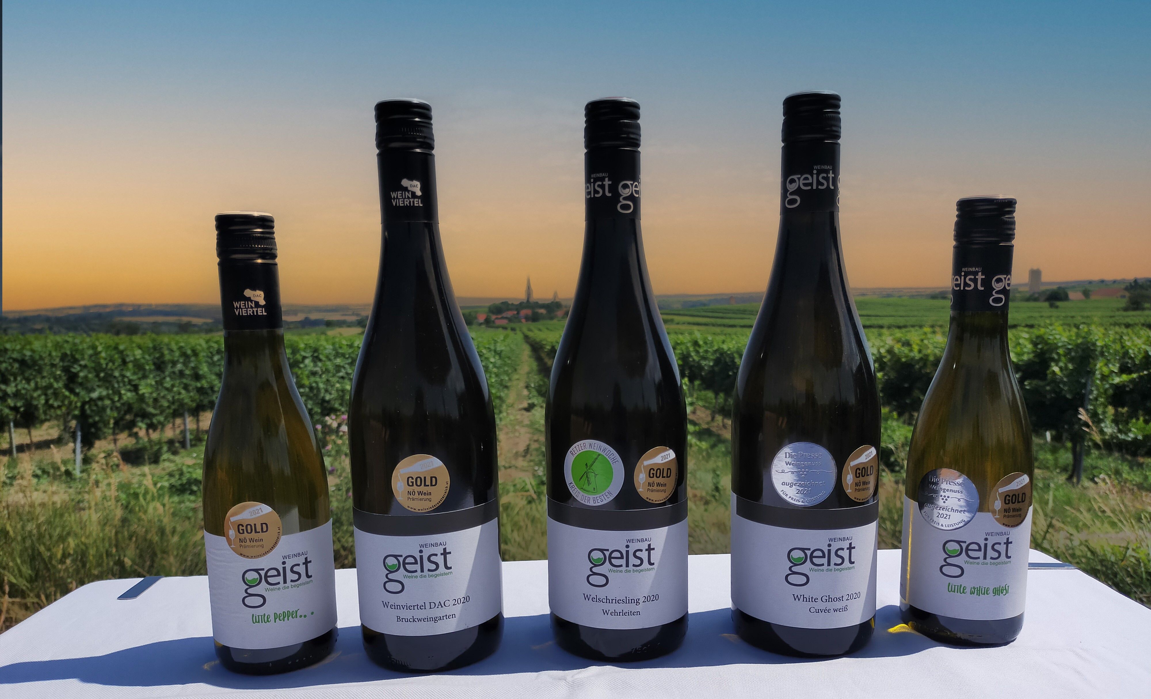 Six Geist wine bottles in front of a vineyard at sunset.