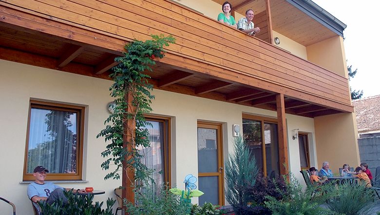 Menschen auf einem Balkon und im Garten eines zweistöckigen Hauses.