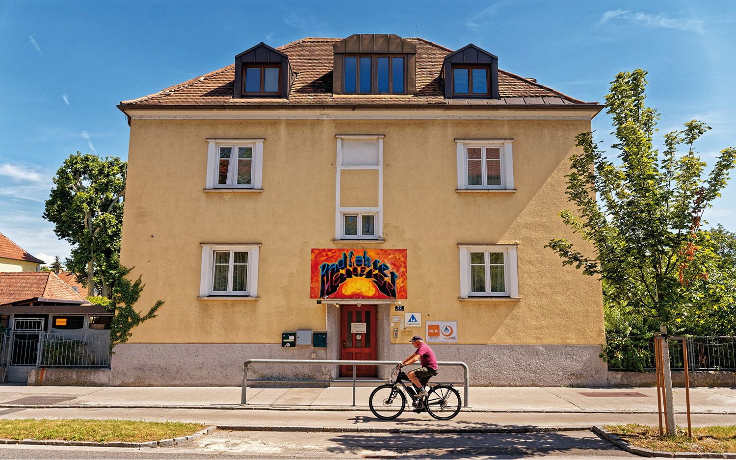 Gelbes Gebäude mit rotem Eingang und Graffiti-Schild. Ein Radfahrer fährt vorbei. Blauer Himmel und Bäume im Hintergrund.