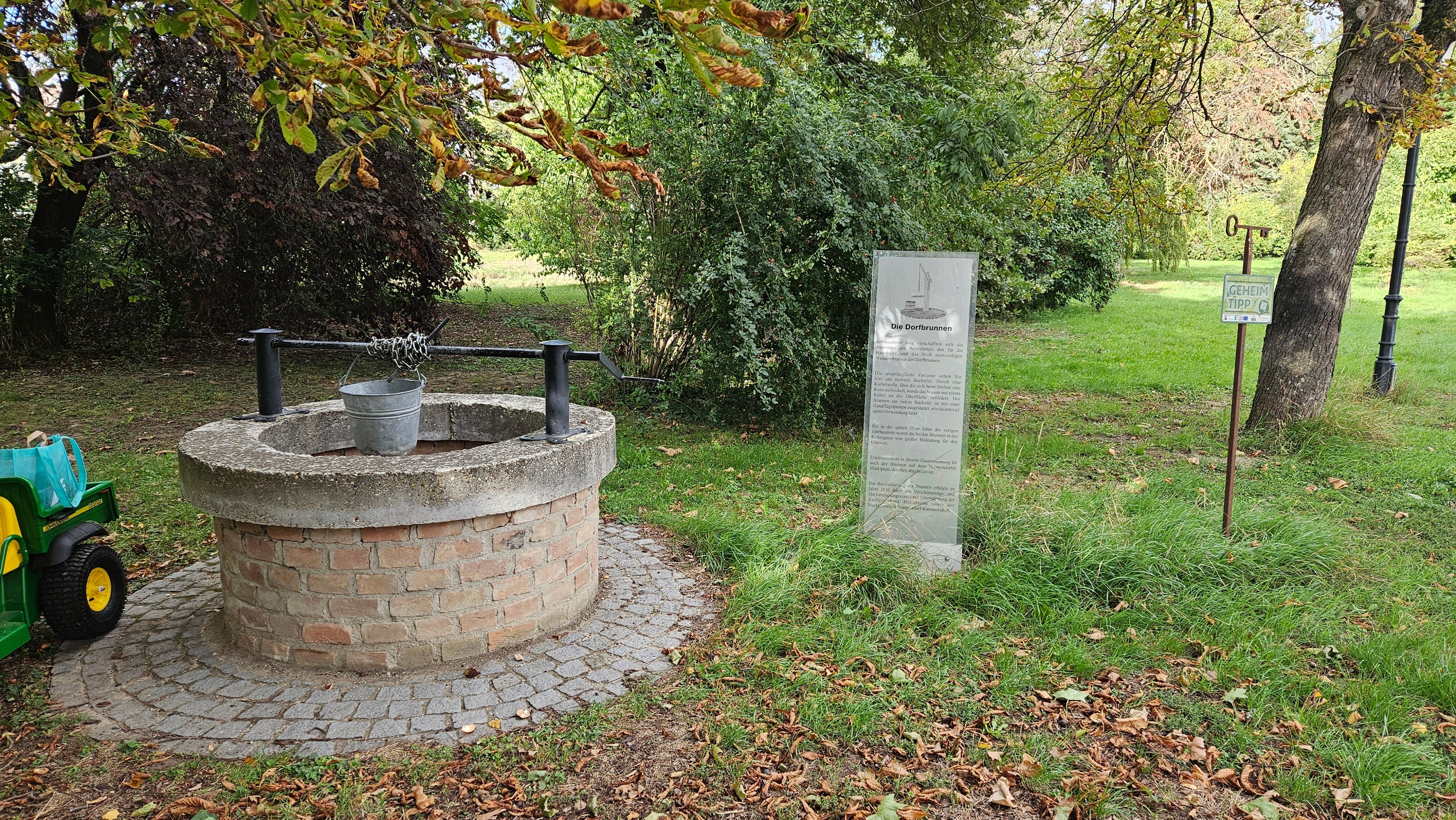 Steinbrunnen in einem grünen Park mit Hinweistafel, Bäumen und einem Schild „Geheimtipp“ in Nappersdorf.