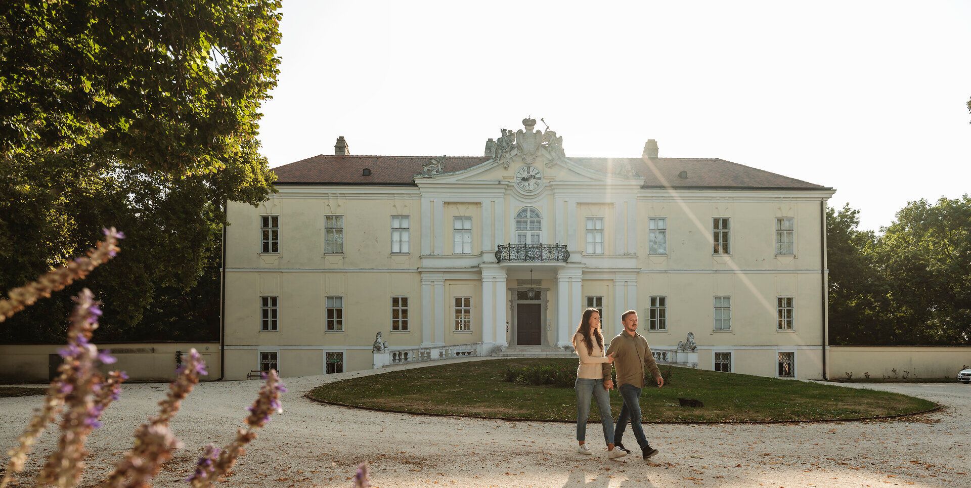 Ein romantischer Spaziergang führt durch die weitläufigen Gärten des Schlosses, wo die sanften Strahlen der Abendsonne die majestätische Fassade erleuchten. Die harmonische Verbindung von Natur und Architektur schafft eine einladende Atmosphäre, die zum Verweilen einlädt.