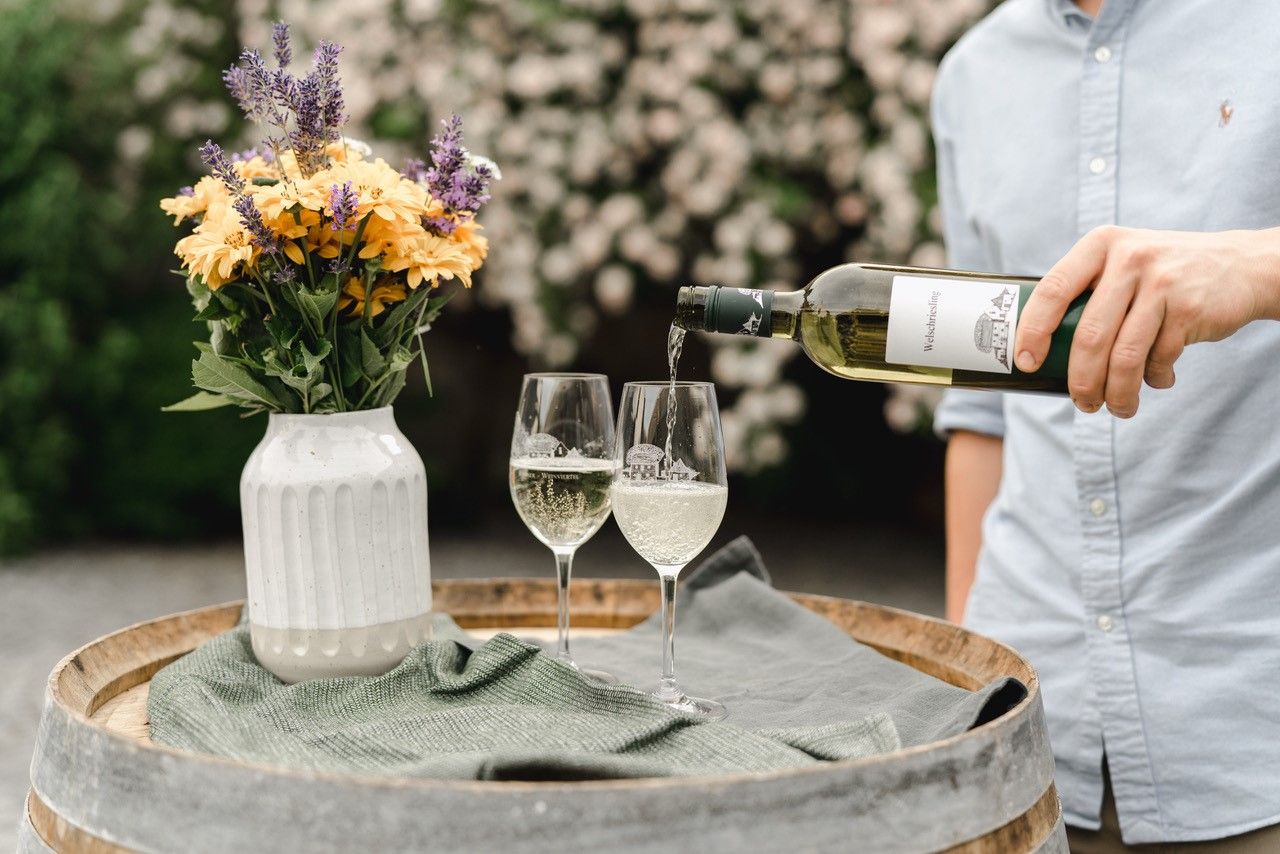 Eine Person schenkt Weißwein in zwei Gläser auf einem Weinfass ein, daneben steht eine Vase mit Blumen.
