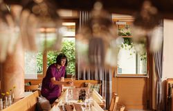 In einem einladenden Restaurant bereitet eine aufmerksame Mitarbeiterin den Tisch für die Gäste vor. Die warmen Holztöne und die frischen Blumen schaffen eine gemütliche Atmosphäre, die zum Verweilen einlädt. Hier wird Kulinarik mit regionalem Flair zelebriert, perfekt für einen genussvollen Abend.