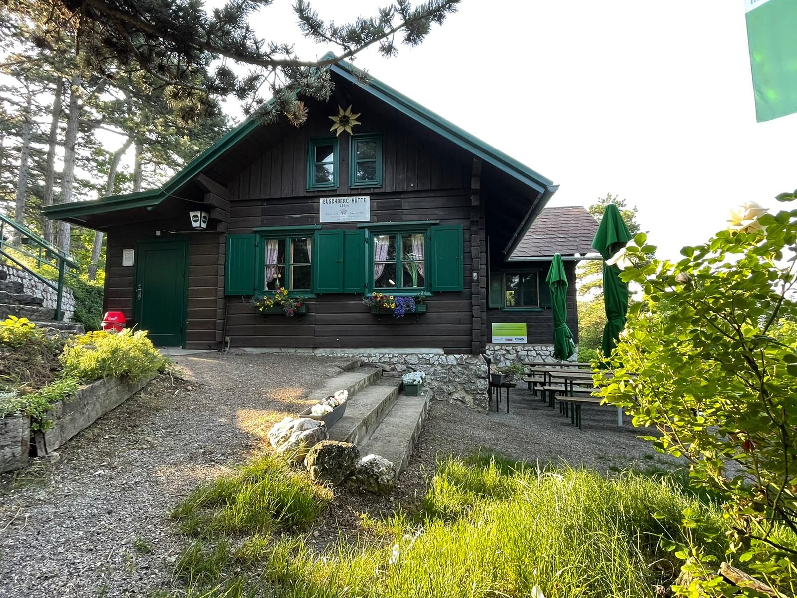 A mountain hut with green shutters and flower boxes, surrounded by trees and garden furniture.