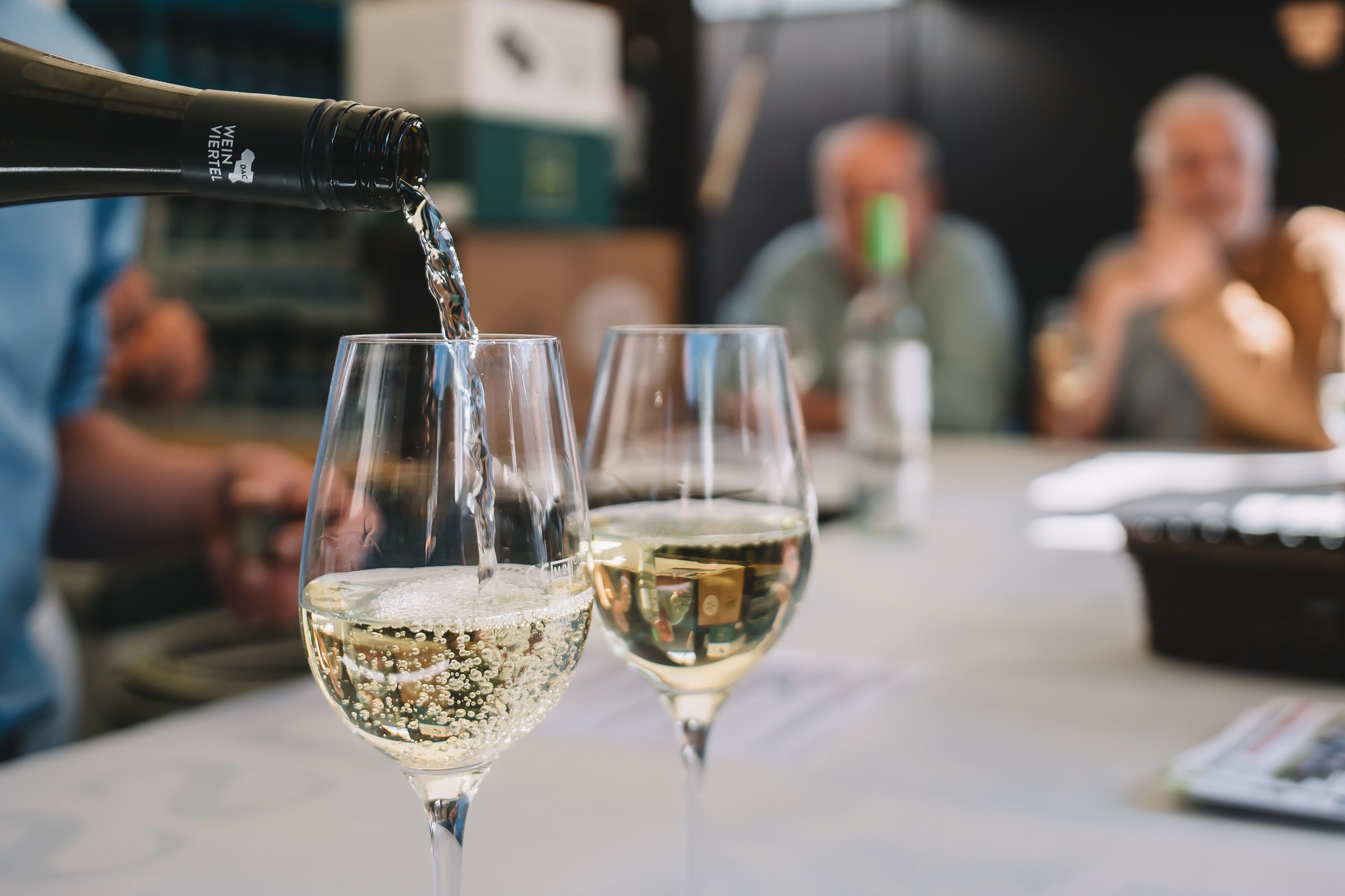Weinviertel DAC Grüner Veltliner wird in ein Weinglas eingeschenkt, daneben steht ein bereits gefülltes Glas. Im Hintergrund sitzen Personen mit einem Weinglas. 