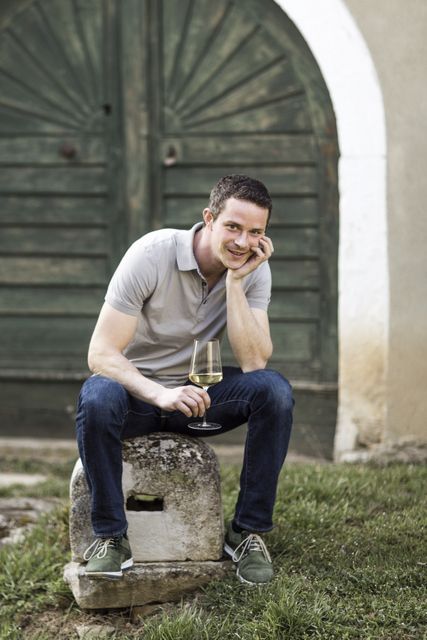 A man sits smiling with a wine glass in his hand in front of an old wooden door.