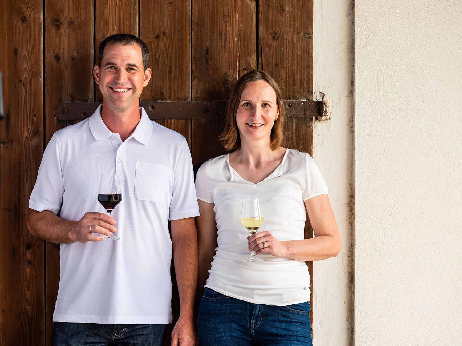 A man and a woman stand smiling in front of a wooden wall, both holding glasses of wine.