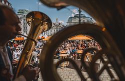 Durch zwei Musikanten, welche auf der Bühne sitzen und musizieren, sieht man Personen, welche Weinviertler Getränke und Kulinarik in einer Innenstadt bei Sonnenschein genießen.