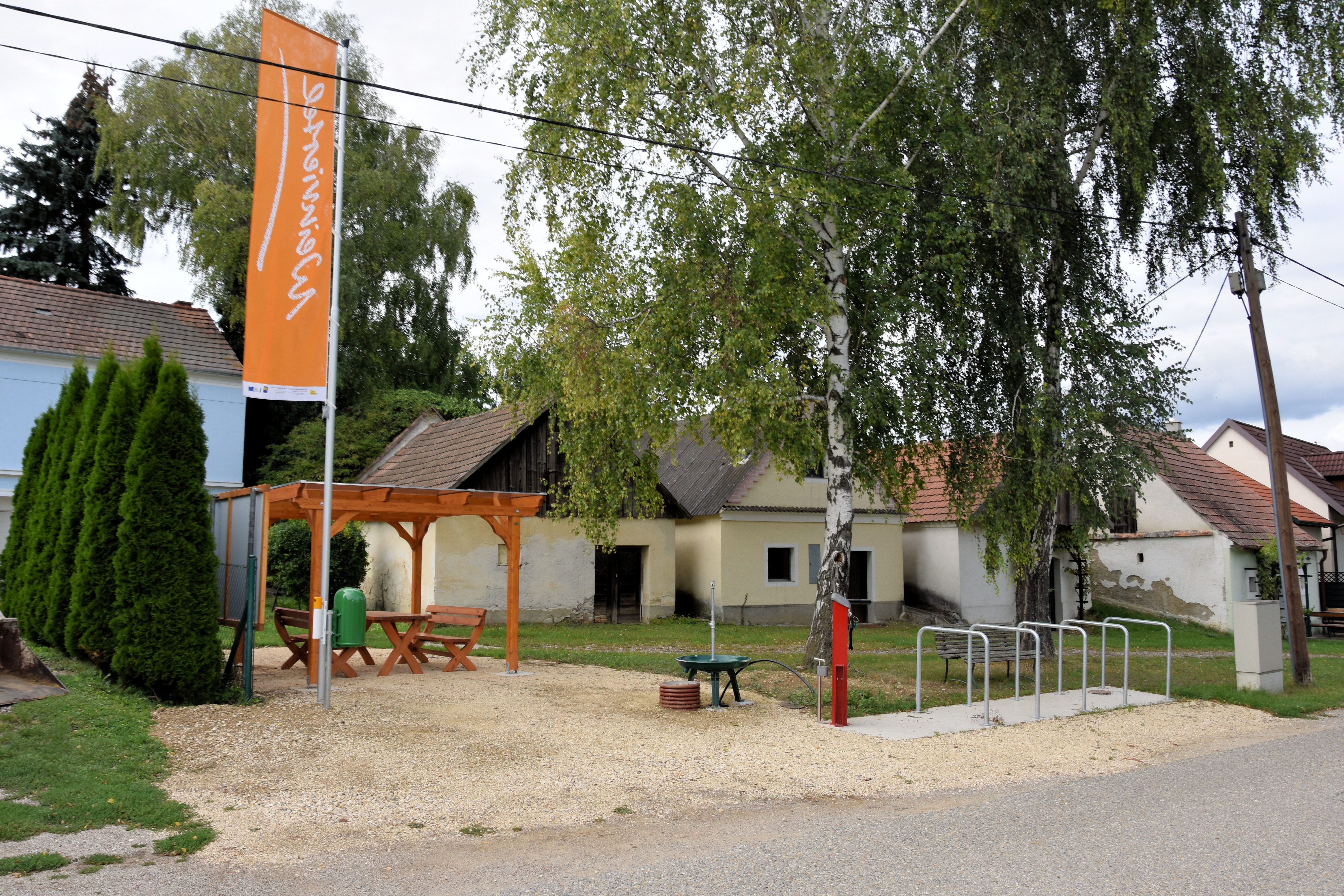 Ein Rastplatz mit Holztisch und Bänken unter einem Pavillon, umgeben von Bäumen und alten Gebäuden. Ein Fahrradständer und eine Infotafel sind ebenfalls vorhanden.