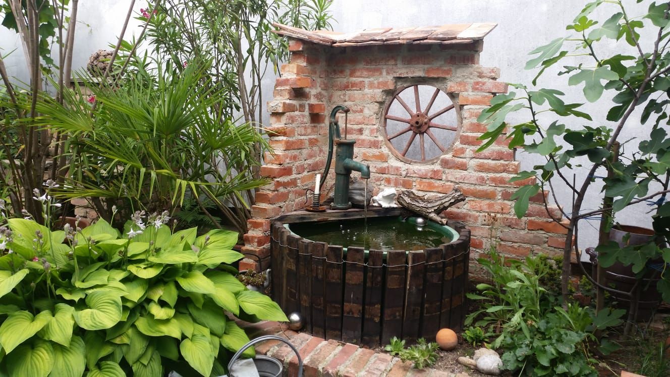 Ein rustikaler Gartenbrunnen mit Backsteinmauer, umgeben von Pflanzen und einem alten Wagenrad als Dekoration.