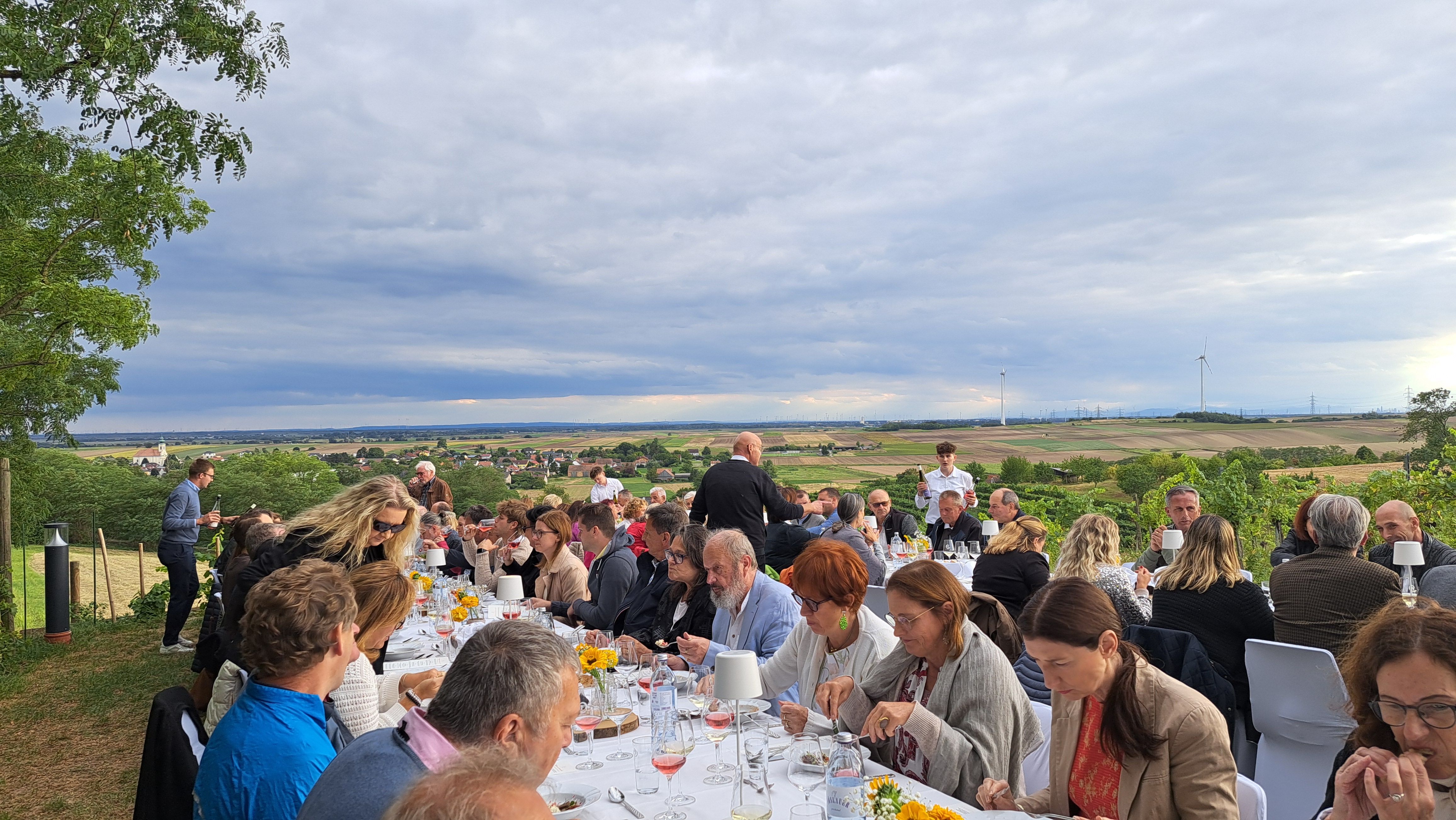 Menschen sitzen an langen Tischen im Freien mit Blick auf eine ländliche Landschaft.