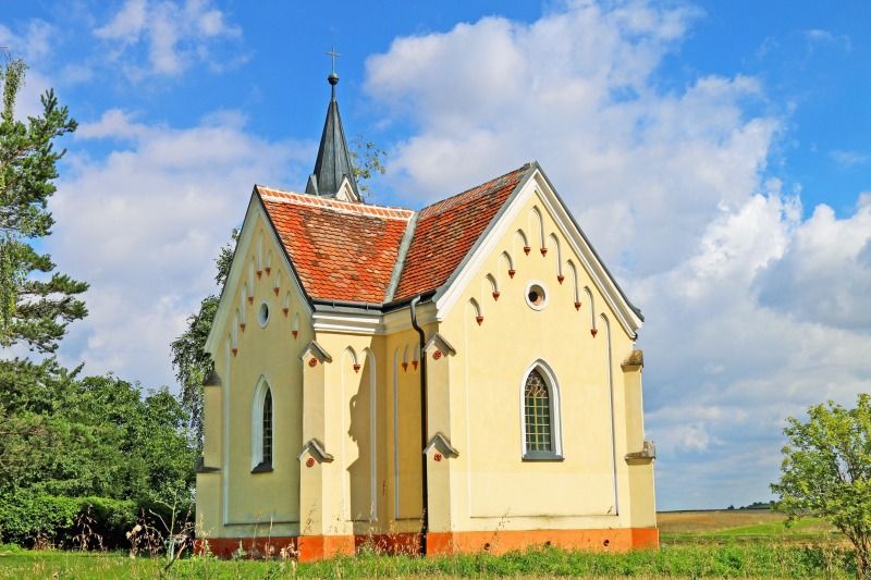 Eine kleine, gelbe Kapelle mit rotem Ziegeldach steht auf einer Wiese unter einem blauen Himmel mit Wolken.