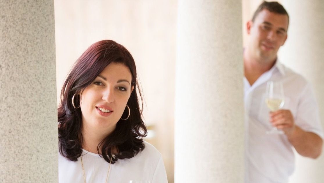 Two people are standing behind pillars, a woman in the foreground and a man in the background with a wine glass.
