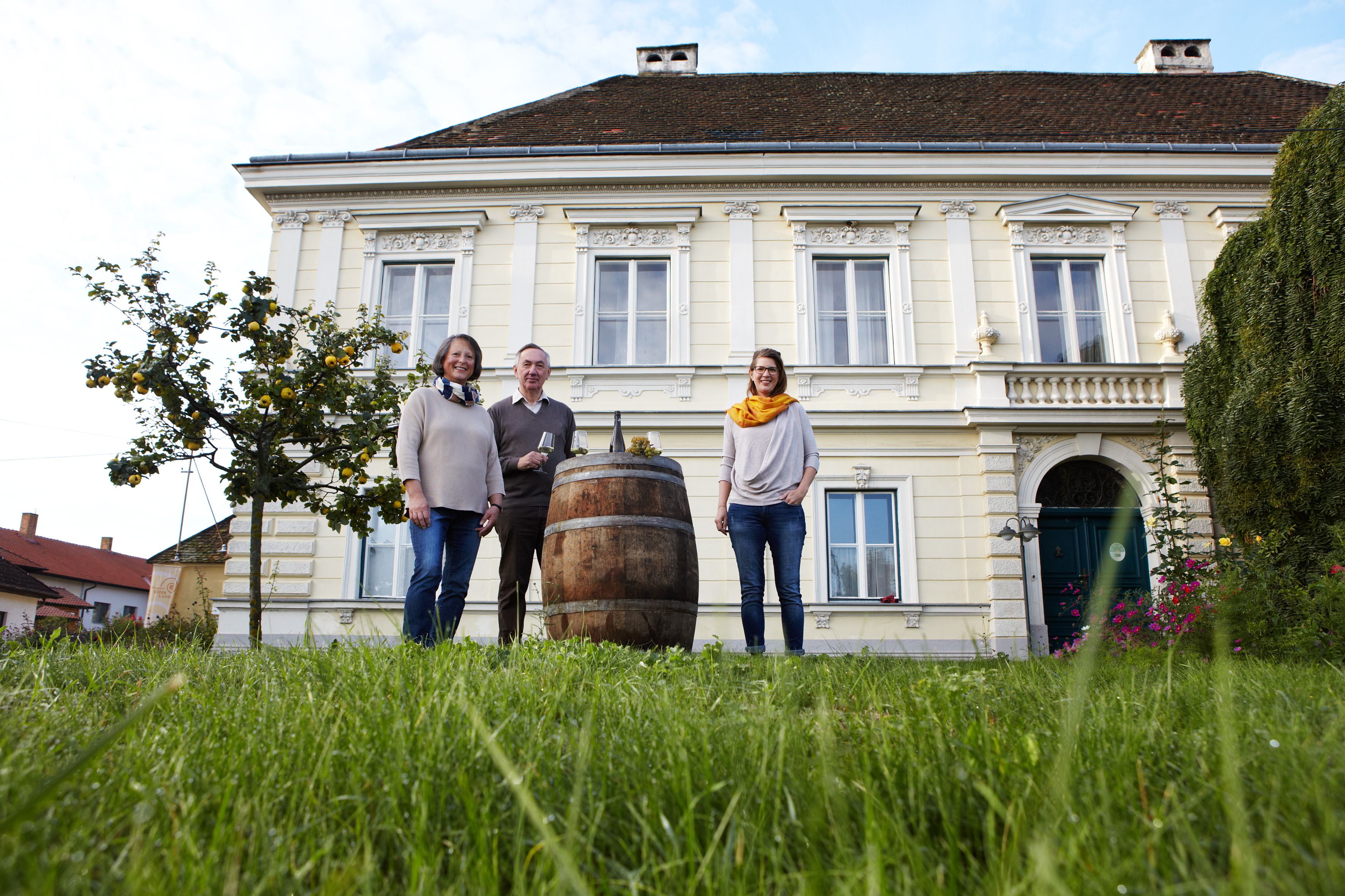 Drei Personen stehen vor einem großen, gelben Gebäude mit einem Fass im Vordergrund.