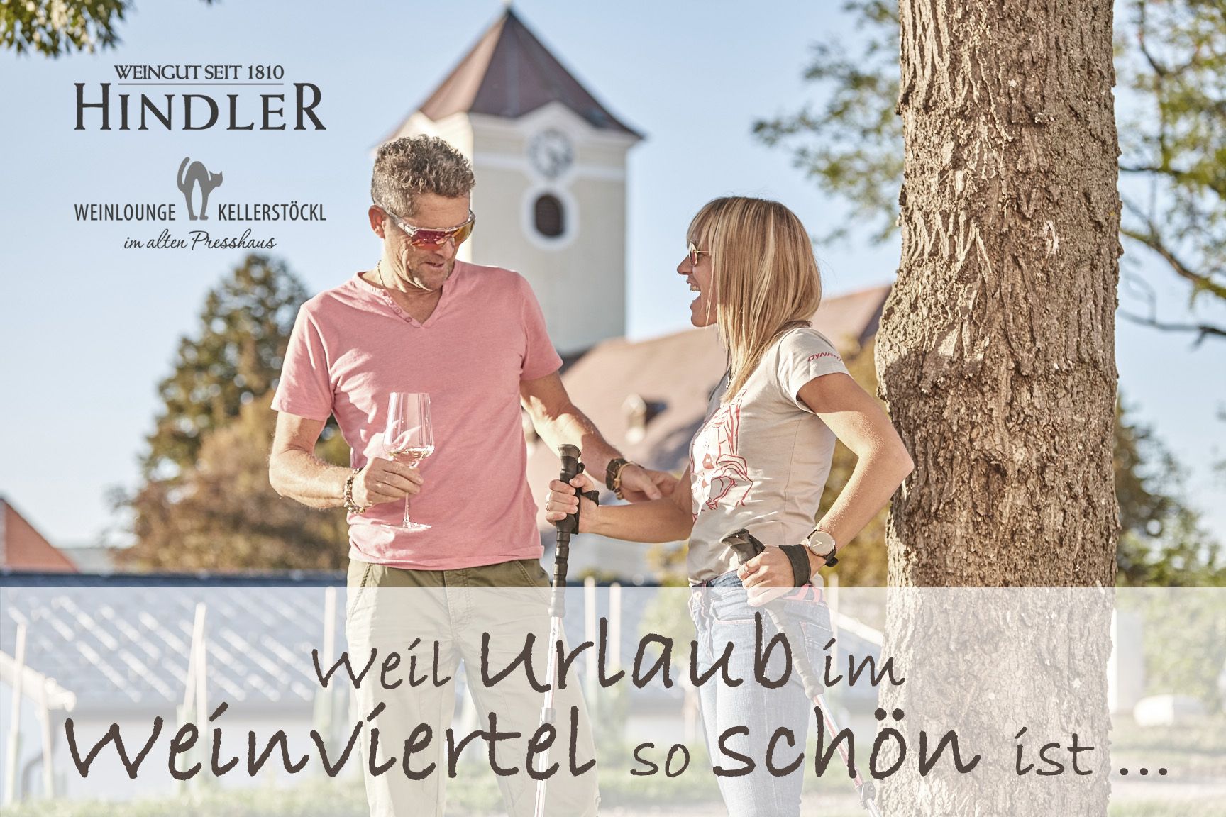 Ein Mann und eine Frau stehen mit Weingläsern und Wanderstöcken vor einem Baum, im Hintergrund ein Gebäude mit Turm.