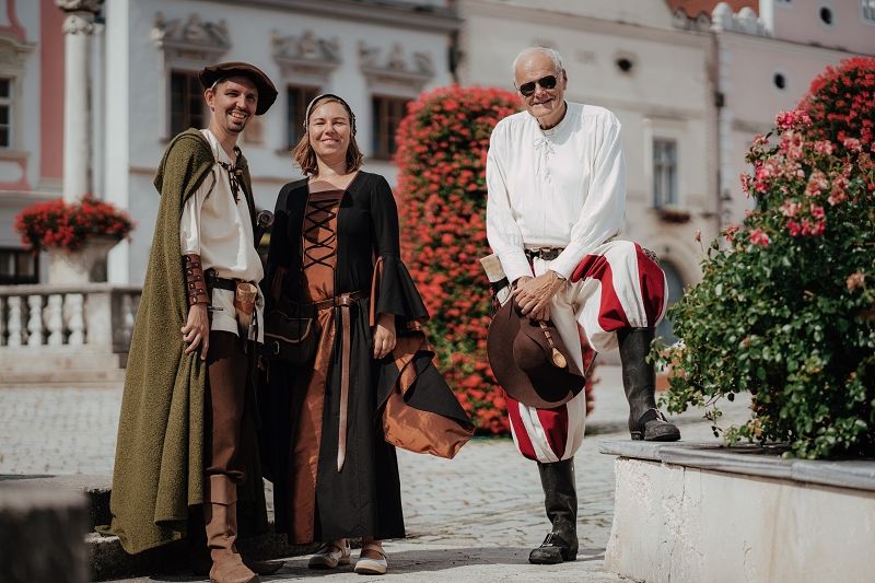 Drei Personen in historischen Kostümen stehen vor einem blumengeschmückten Platz.