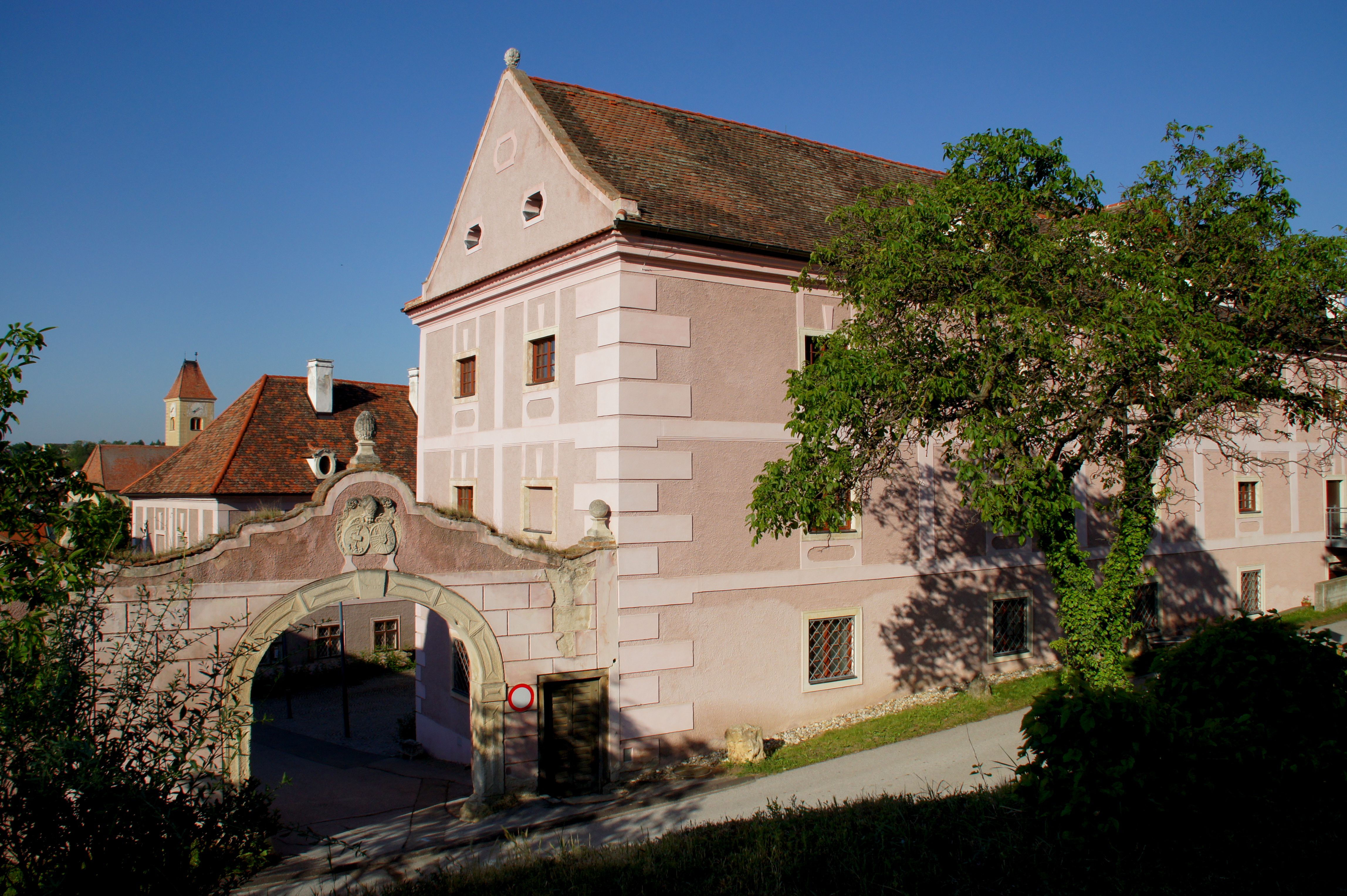 Historisches Gebäude mit rosa Fassade und Torbogen, umgeben von Bäumen.