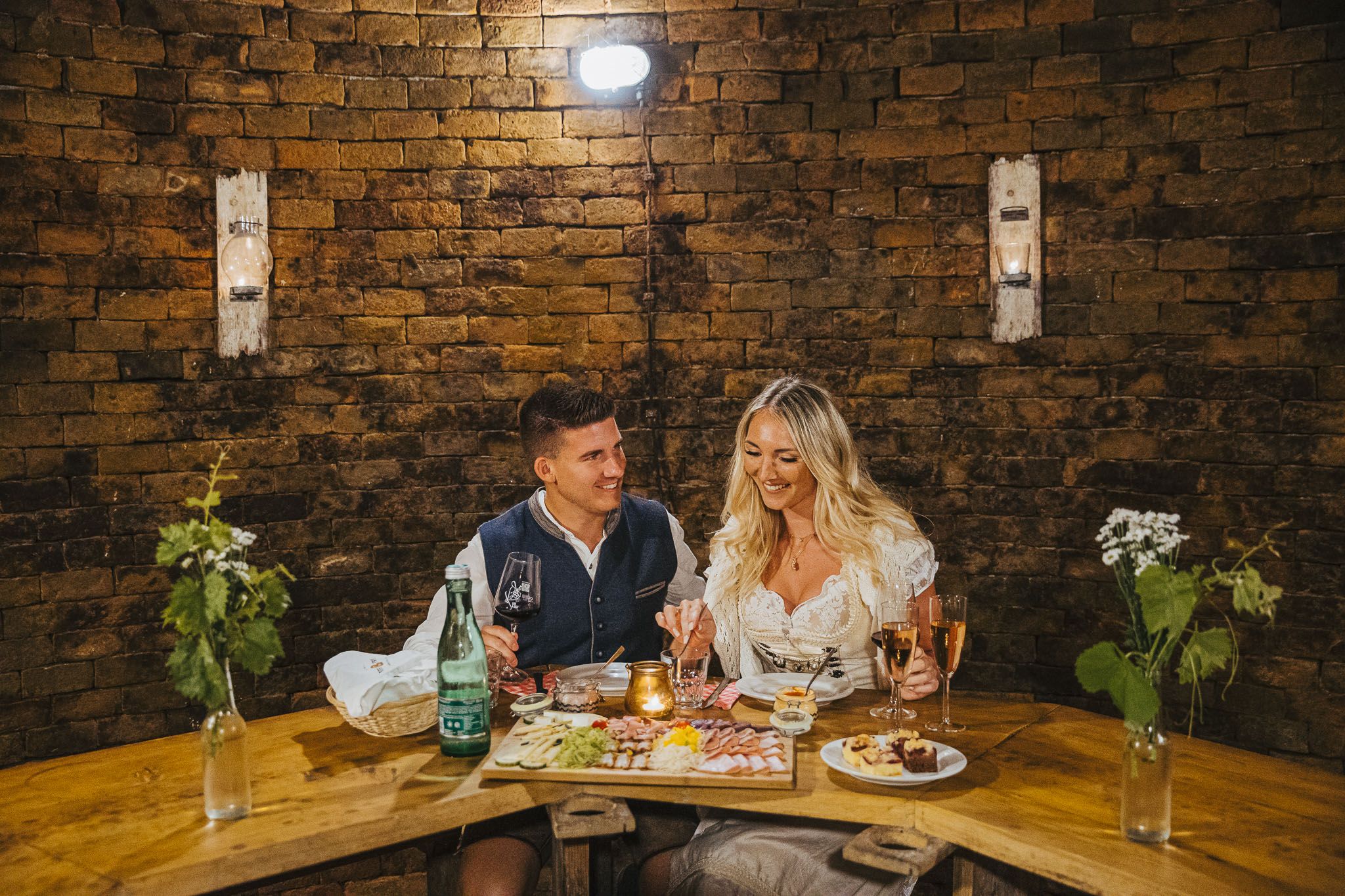 Paar beim Essen im Winzerhof Küssler an einem rustikalen Holztisch mit Wein und Speisen vor einer Backsteinwand.
