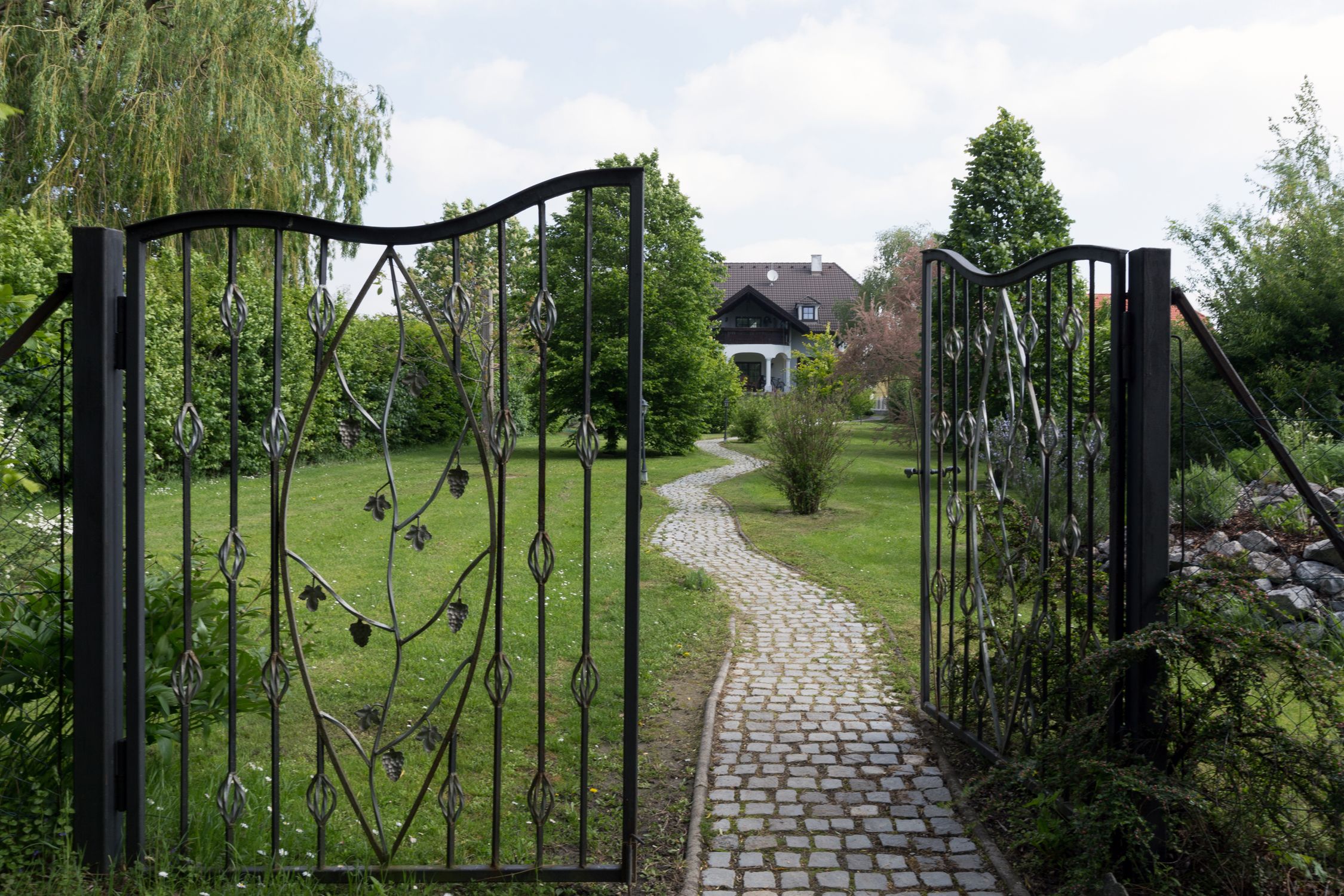 Ein offenes schmiedeeisernes Tor führt zu einem gepflasterten Weg, der durch einen grünen Garten zu einem Haus im Hintergrund führt.