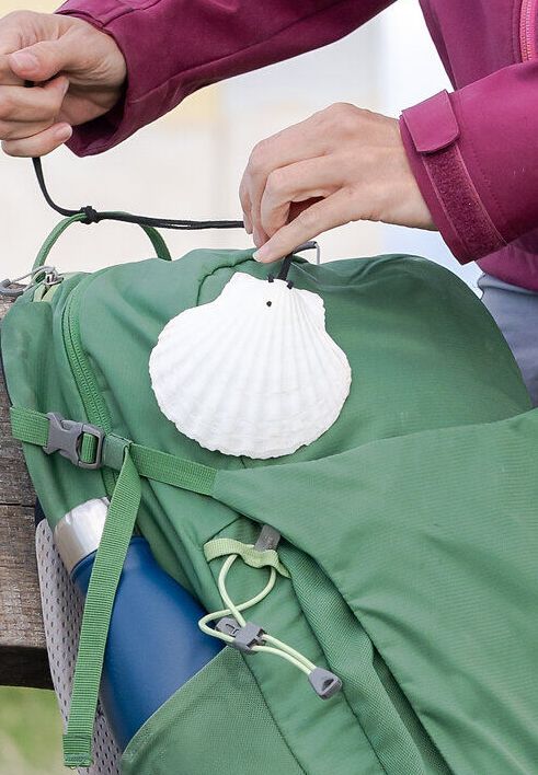 Eine Person befestigt eine Jakobsmuschel auf einem Wanderrucksack mit Trinkflasche, der auf einer Holzbank abgestellt ist.