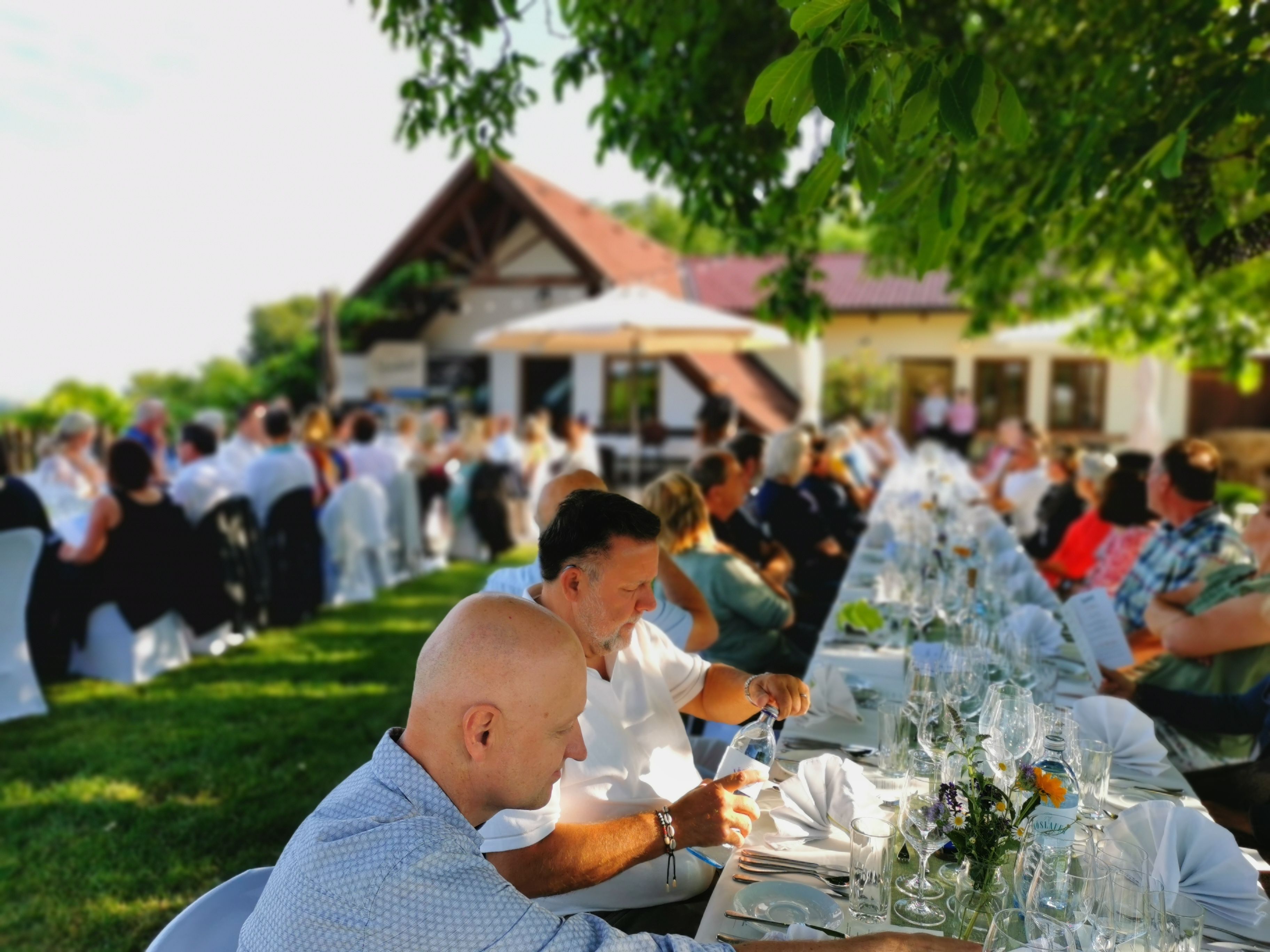 Menschen sitzen an langen, gedeckten Tischen im Freien bei einer Veranstaltung.