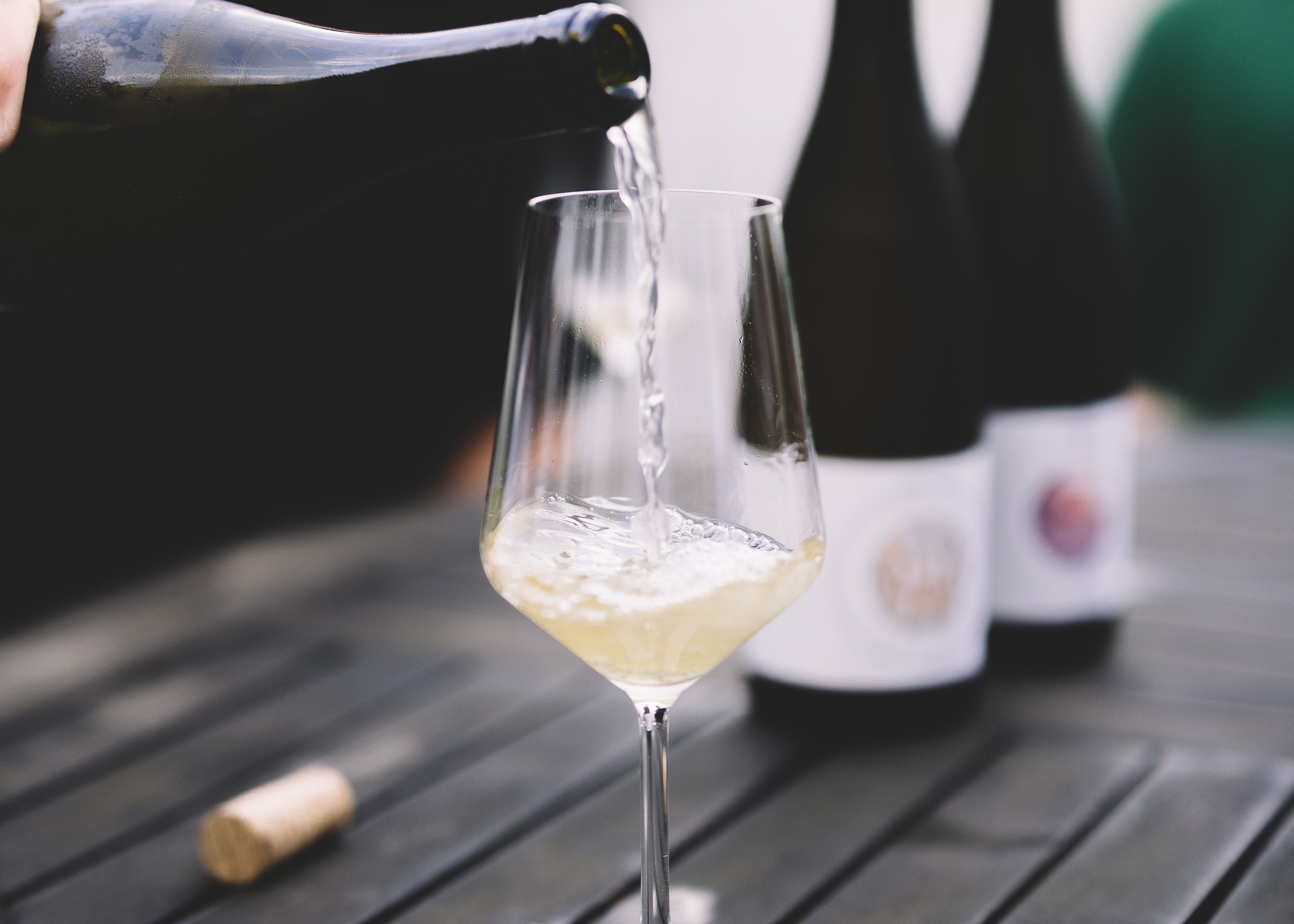 White wine is poured into a glass on a wooden table, with two bottles of wine in the background.