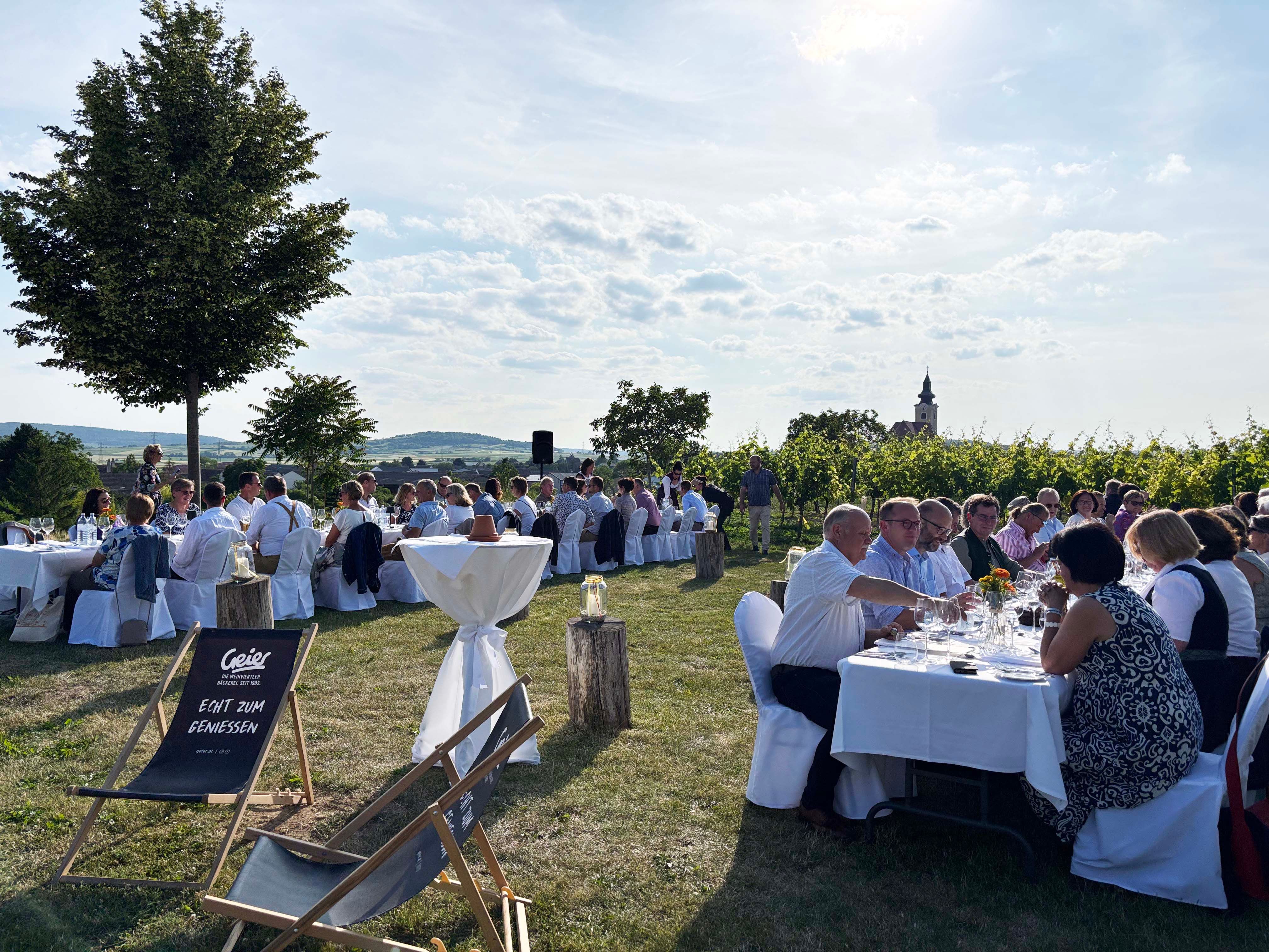 Menschen sitzen an gedeckten Tischen im Freien, umgeben von Weinbergen und Bäumen, mit einer Kirche im Hintergrund.