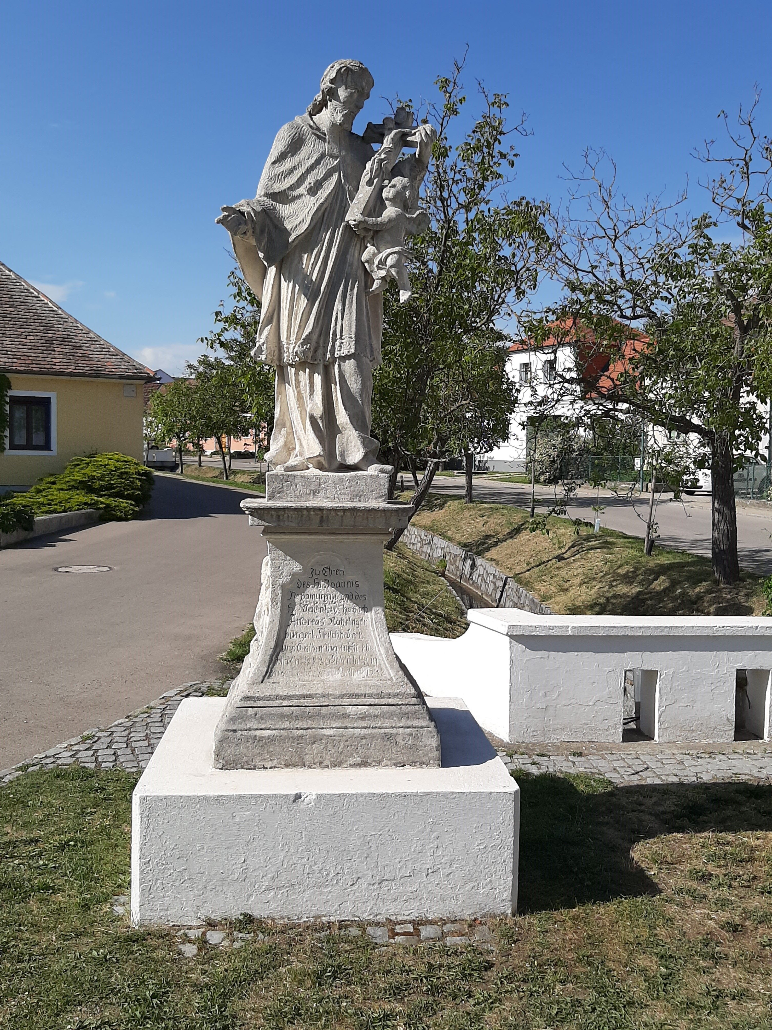 Statue des Heiligen Johannes Nepomuk in Obermarkersdorf, umgeben von Bäumen und Gebäuden.