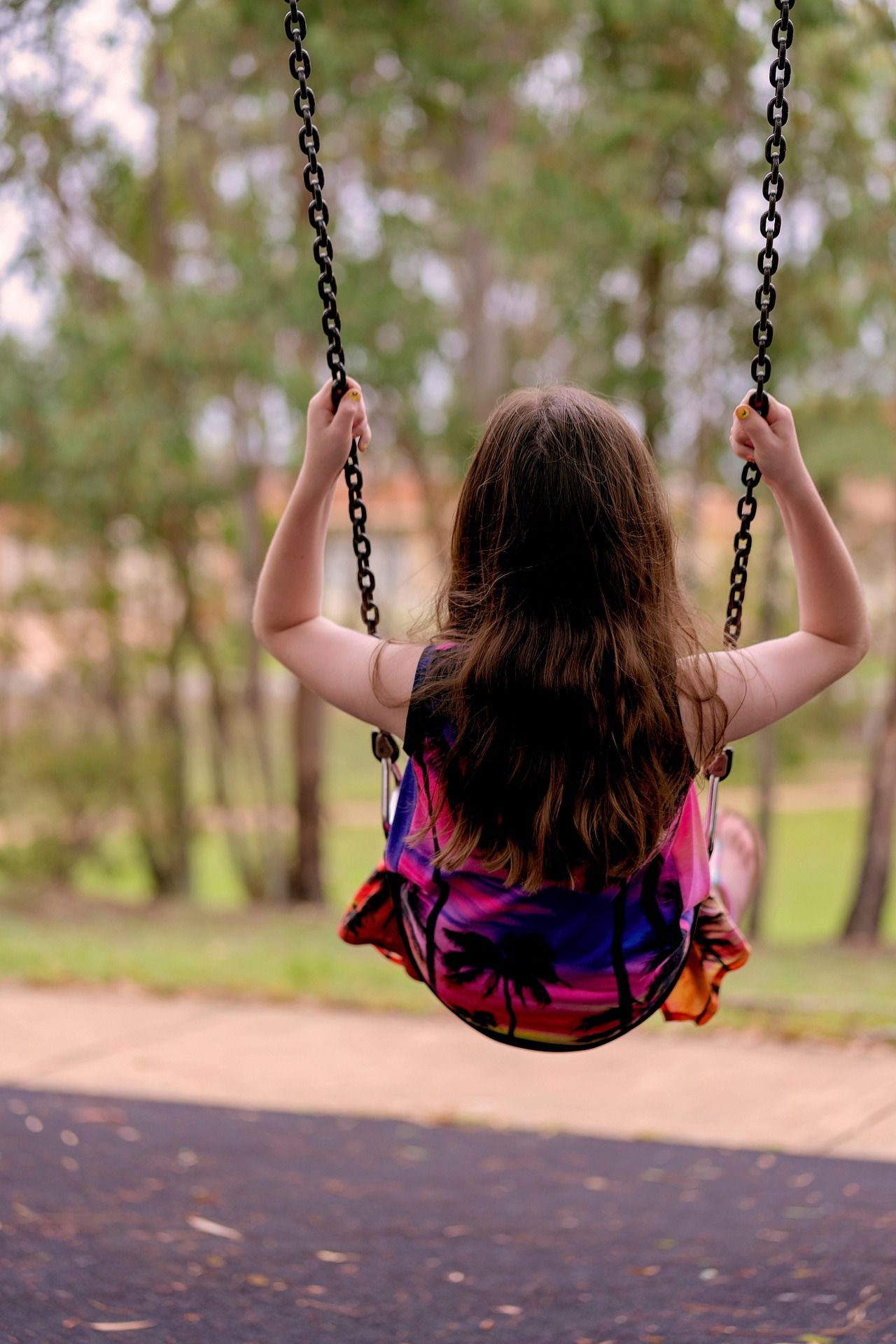 A child swings outdoors, photographed from behind, surrounded by trees.