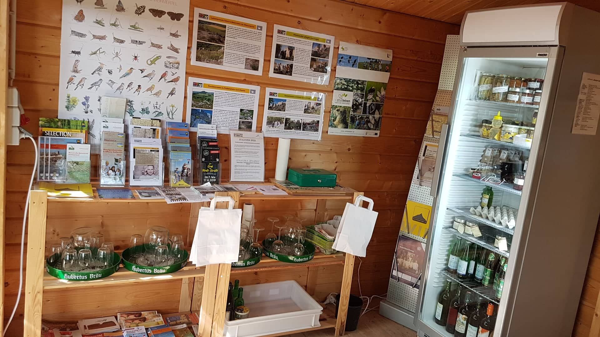 Innenansicht eines kleinen Verkaufsstands mit Infomaterial und Kühlschrank.