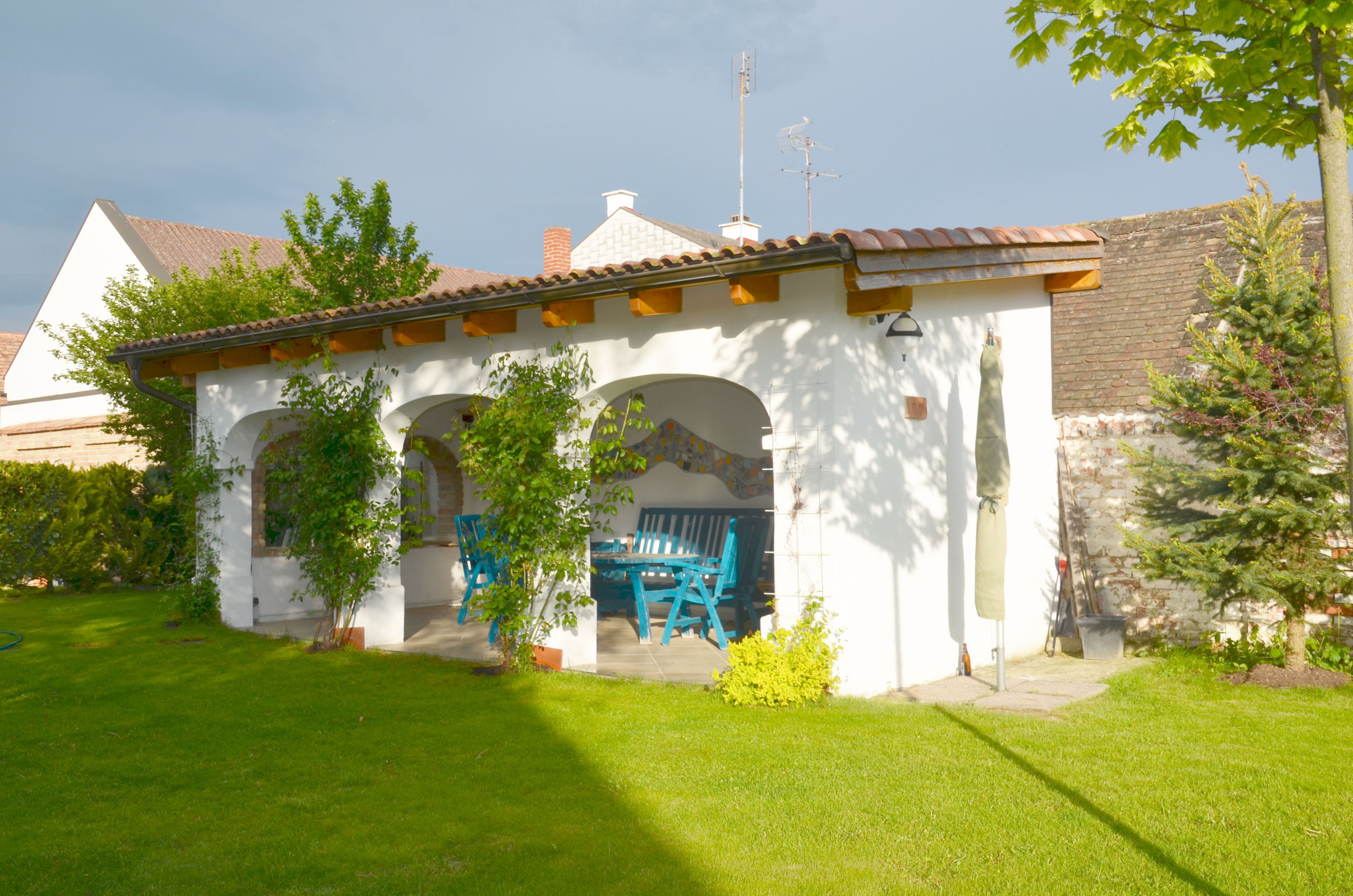 A garden with a covered terrace, blue furniture and green vegetation.