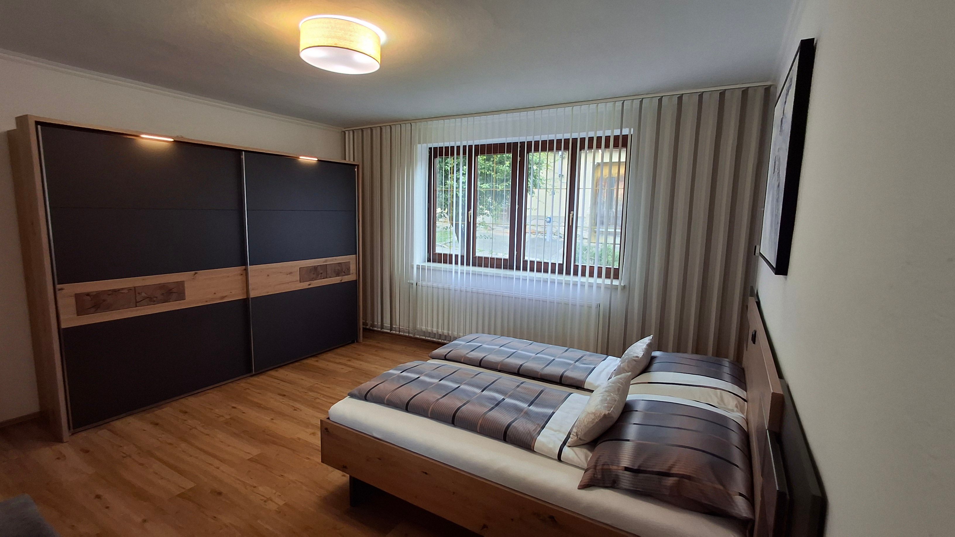 Modernes Schlafzimmer mit Doppelbett, großem Kleiderschrank und Fenster mit Jalousien.