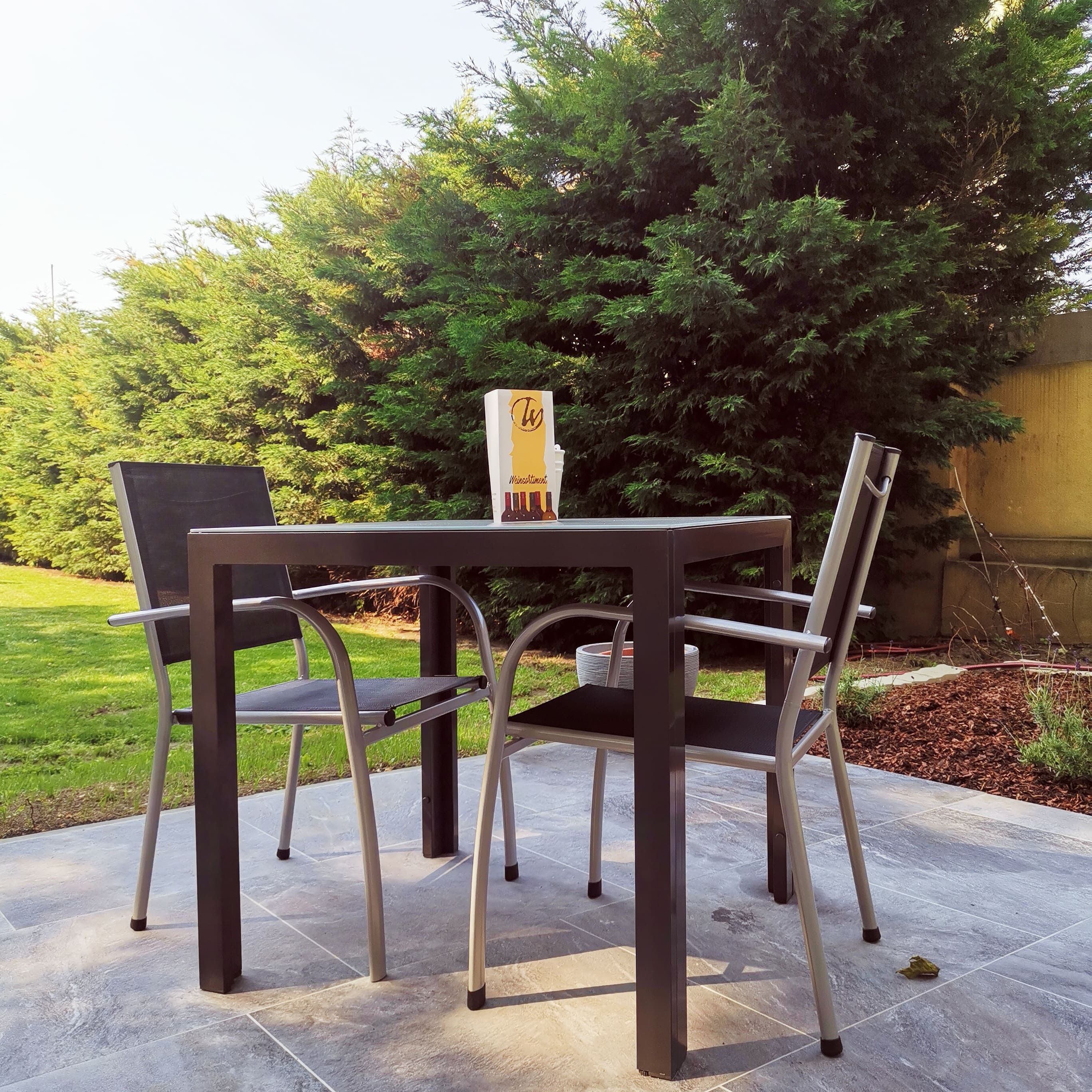 Terrasse mit Tisch, zwei Stühlen und Bäumen im Hintergrund.