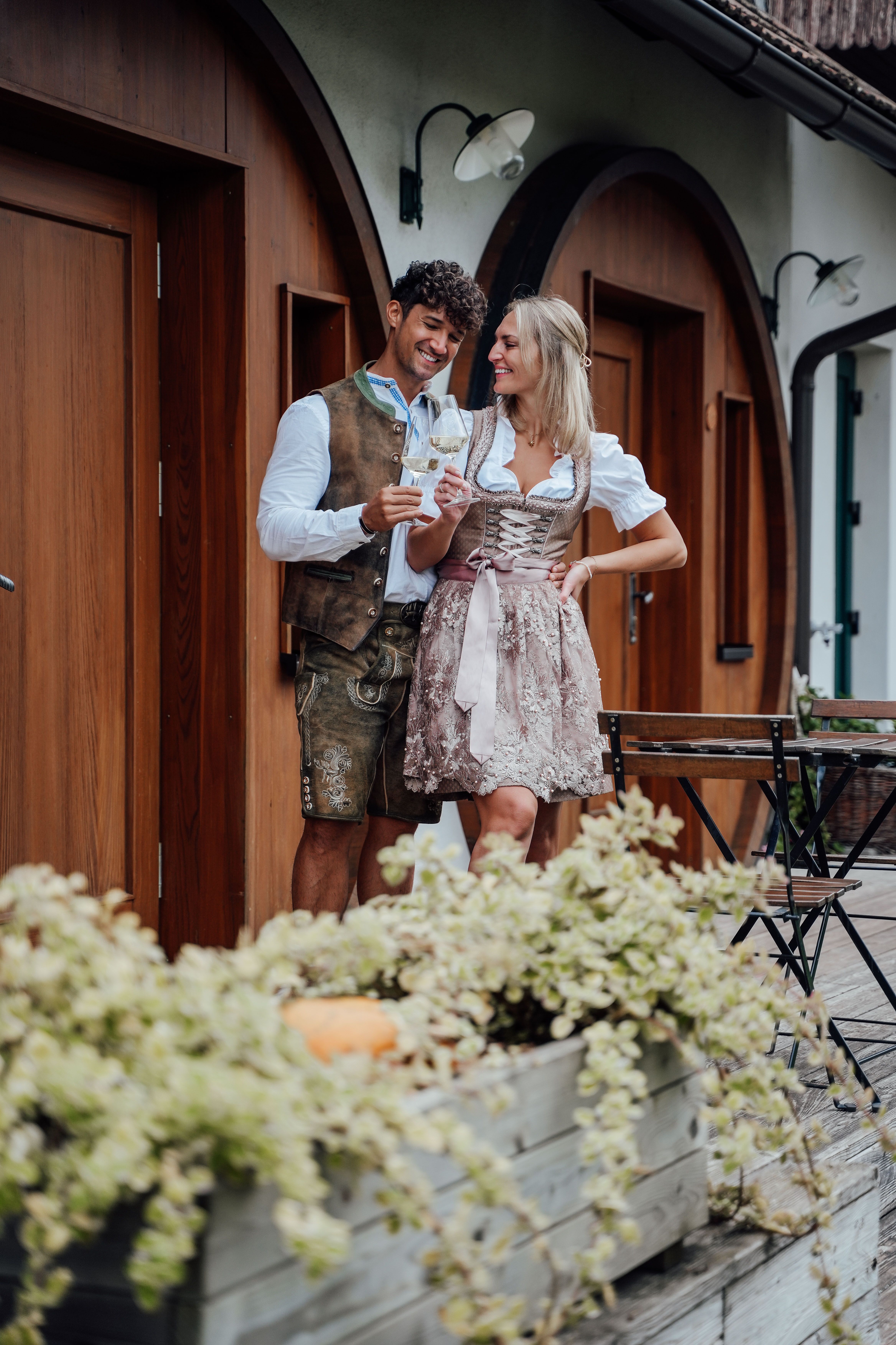 Paar in Tracht stößt mit Weißweingläsern vor dem Winzerhof Küssler an, vor einer Holzfassade mit Rundbögen und Blumenkästen.
