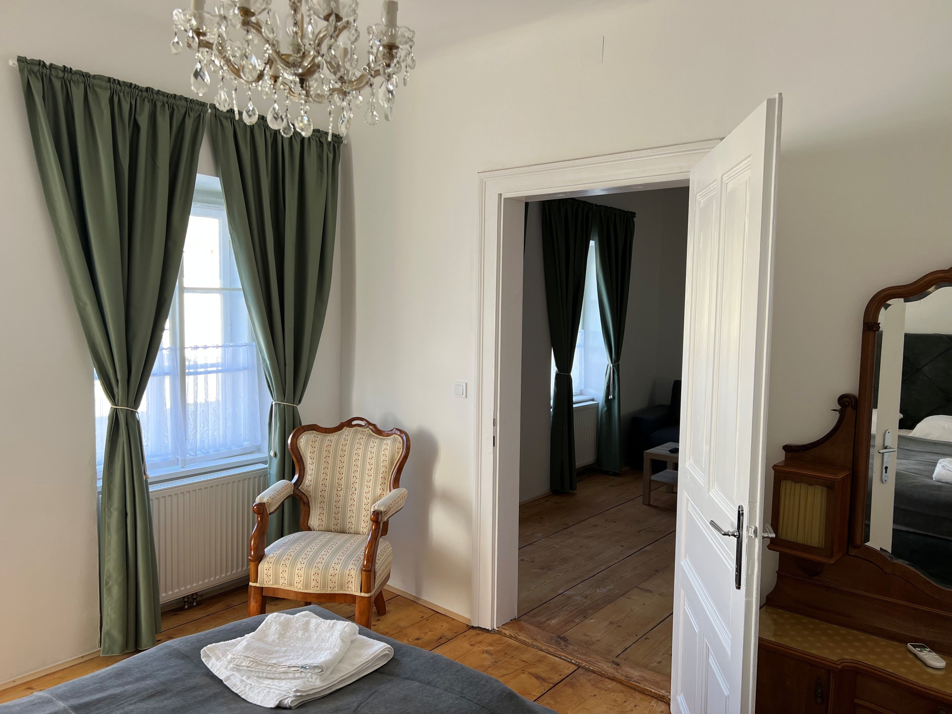Ein elegantes Schlafzimmer mit einem antiken Sessel, grünen Vorhängen und einem Kronleuchter. Eine offene Tür führt in einen angrenzenden Raum.