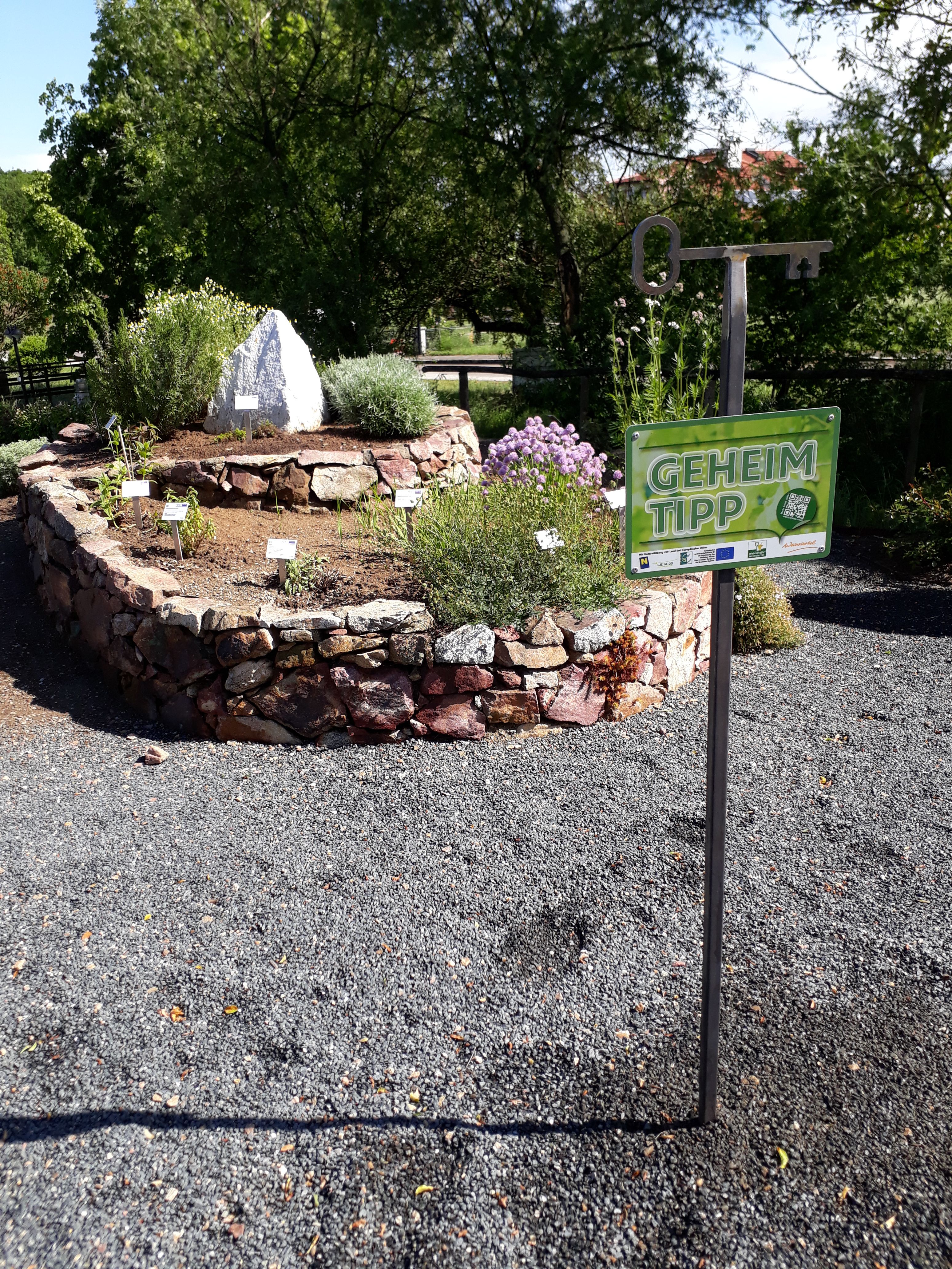 Kräuterspirale aus Steinen mit Pflanzen und einem Schild mit der Aufschrift 'Geheimtipp'.